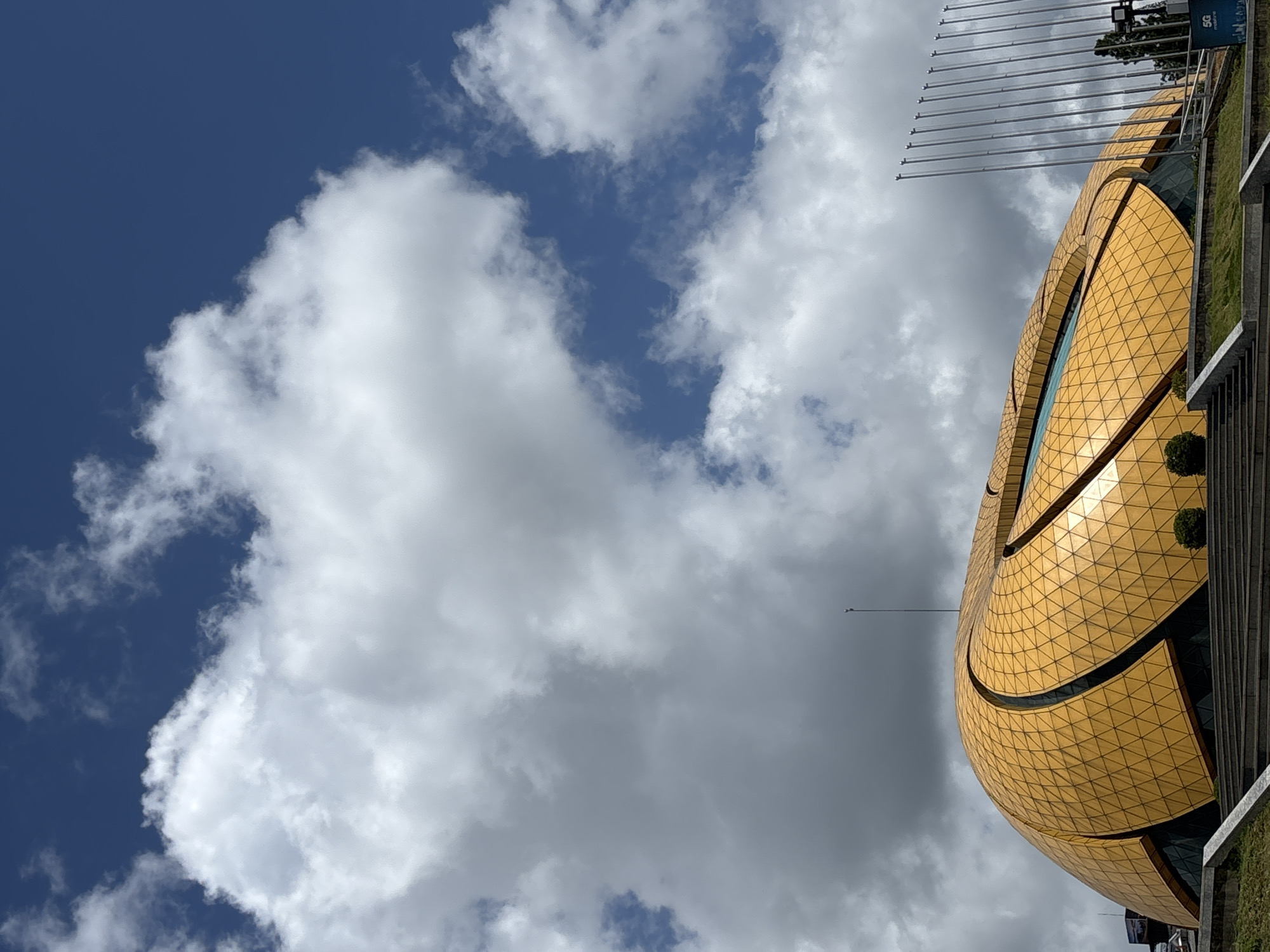 A low-angle, full-shot photograph captures the iconic golden dome of the Da Lat Flower Park Convention Center, also known as the Da Lat Cultural and Conference Center, against a vibrant blue sky filled with dramatic white clouds. The building's unique, lotus-shaped architecture is highlighted by its golden, tessellated facade, which reflects the sunlight. The structure appears to be empty, with no people visible in the frame. In the foreground, stone steps lead up to the building, bordered by lush green grass. To the right of the convention center, a row of tall, slender flagpoles stands silhouetted against the sky. The overall scene conveys a sense of grandeur and architectural brilliance within a bright, open environment.