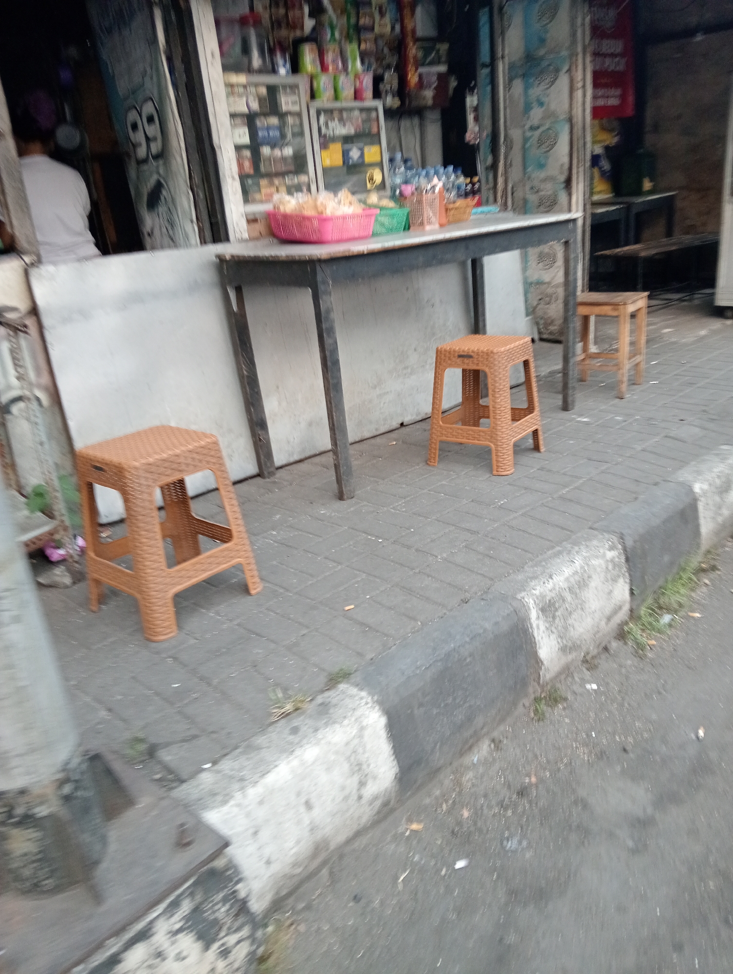 The image depicts a street scene in Sinduadi, Indonesia, likely in front of a small shop. In the foreground, there are two brown plastic stools on a paved sidewalk. One stool is closer to the viewer, and another is further back. A table with a dark metal frame and a lighter top stands behind the first stool, extending towards the shop entrance. On the table, a pink basket filled with what appears to be snacks is visible, along with other items that are difficult to discern due to the image quality. Behind the table, the shop entrance is visible, showing shelves stocked with various products, including drinks and packaged goods. A person in a white shirt is partially visible inside the shop. The sidewalk is made of grey square tiles, and a concrete curb separates it from the asphalt road, which has some litter on it, including what appear to be cigarette butts. The overall environment suggests a commercial area with outdoor seating for customers.