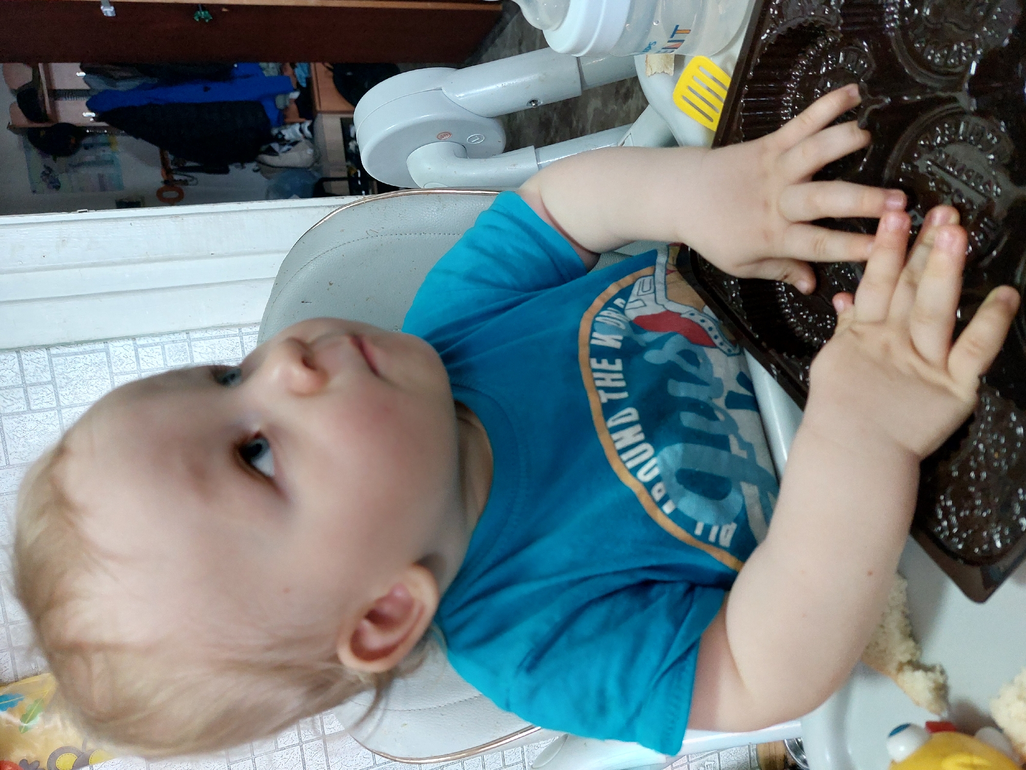 A young child with blond hair and blue eyes sits in a high chair. The child is wearing a blue t-shirt. The child is touching a dark brown plastic baking pan with their hands. A bottle and a yellow spatula are also visible on the high chair tray. The background shows a white tiled floor and a partially visible dresser with clothes. The location is stated to be in the city of Chaikovsky, Russia.