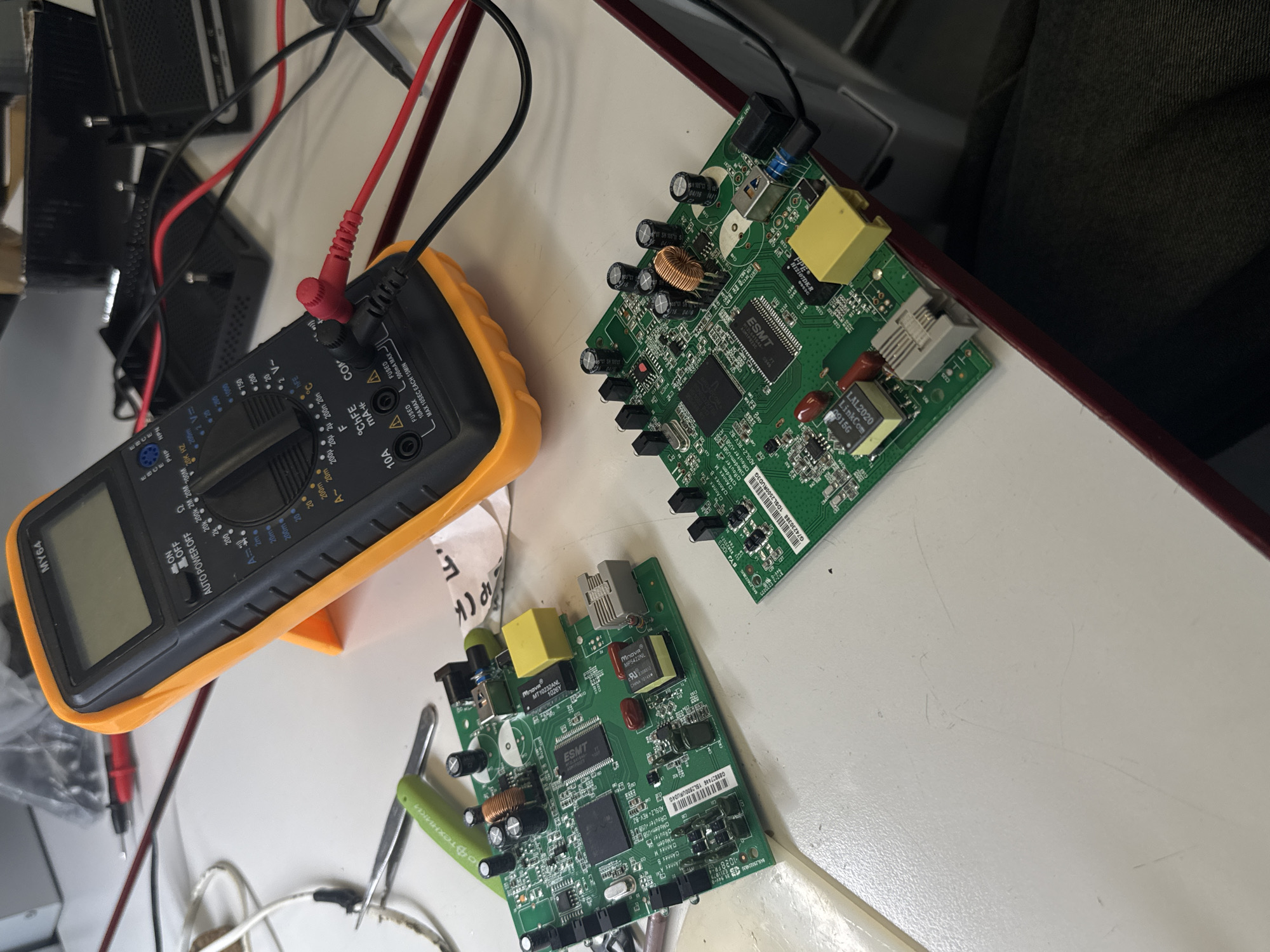 The image captures a technical workspace, likely an electronics repair or testing bench, set in Perm, Russia. The main subjects are a yellow and black digital multimeter and two identical green circuit boards.
The multimeter, model MY64, rests on a stand with its red and black probes connected. Its dial indicates readiness for various electrical measurements including AC/DC voltage and current, resistance, capacitance, temperature, and hFE. Notable labels on the device include "ON," "OFF," "AUTO POWER OFF," "COMM," "VΩmA+," "10A MAX," and warnings about fused inputs.
Two green printed circuit boards (PCBs) are displayed on the white desk. Each board is densely populated with electronic components such as integrated circuits (ICs), electrolytic capacitors, inductors, transformers (e.g., "LinkCom LAL2020 09156"), and various ports. Visible markings on the ICs include "ESMT," "Mnova MT10232ANL 1026Y," and "Mnova MP5422NL." Stickers on the PCBs identify them as "ADSL2+ REV-B2 Router Modem USB" boards, with specific model numbers like "G88827446 15L2500URUD4G" and "G74230388 1DSL2500RUD3G." A date code "04/16 14/1" is visible on one board.
Other objects present on the desk include a pair of green-handled tweezers, various cables, and a crumpled piece of paper or plastic with partial Russian text "офтехники" (likely "of technicians" or "tech specialists") and "PCK". In the background, additional electronic equipment and tangled cables are visible, confirming the technical environment. The setup suggests an ongoing activity of inspection, diagnosis, or repair of the ADSL modem/router boards. The scene is illuminated by artificial indoor lighting.