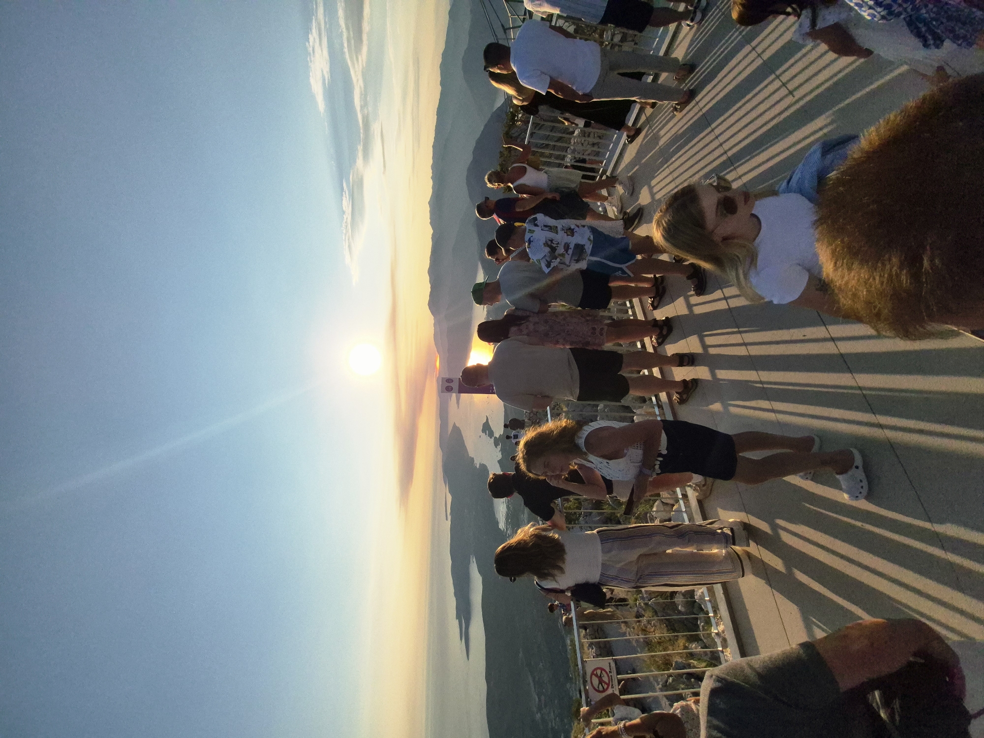 A group of people stand on a viewing platform overlooking a bay at sunset. The sun is low on the horizon, casting a warm glow over the water and surrounding mountains. The platform has a metal railing, and some people are leaning on it, looking out at the view. The shadows of the people and the railing stretch across the paved surface of the platform. The sky is a soft blue, with a few wispy clouds. In the distance, mountains can be seen, and the water is a deep blue. The image was taken in Kuk, Montenegro.