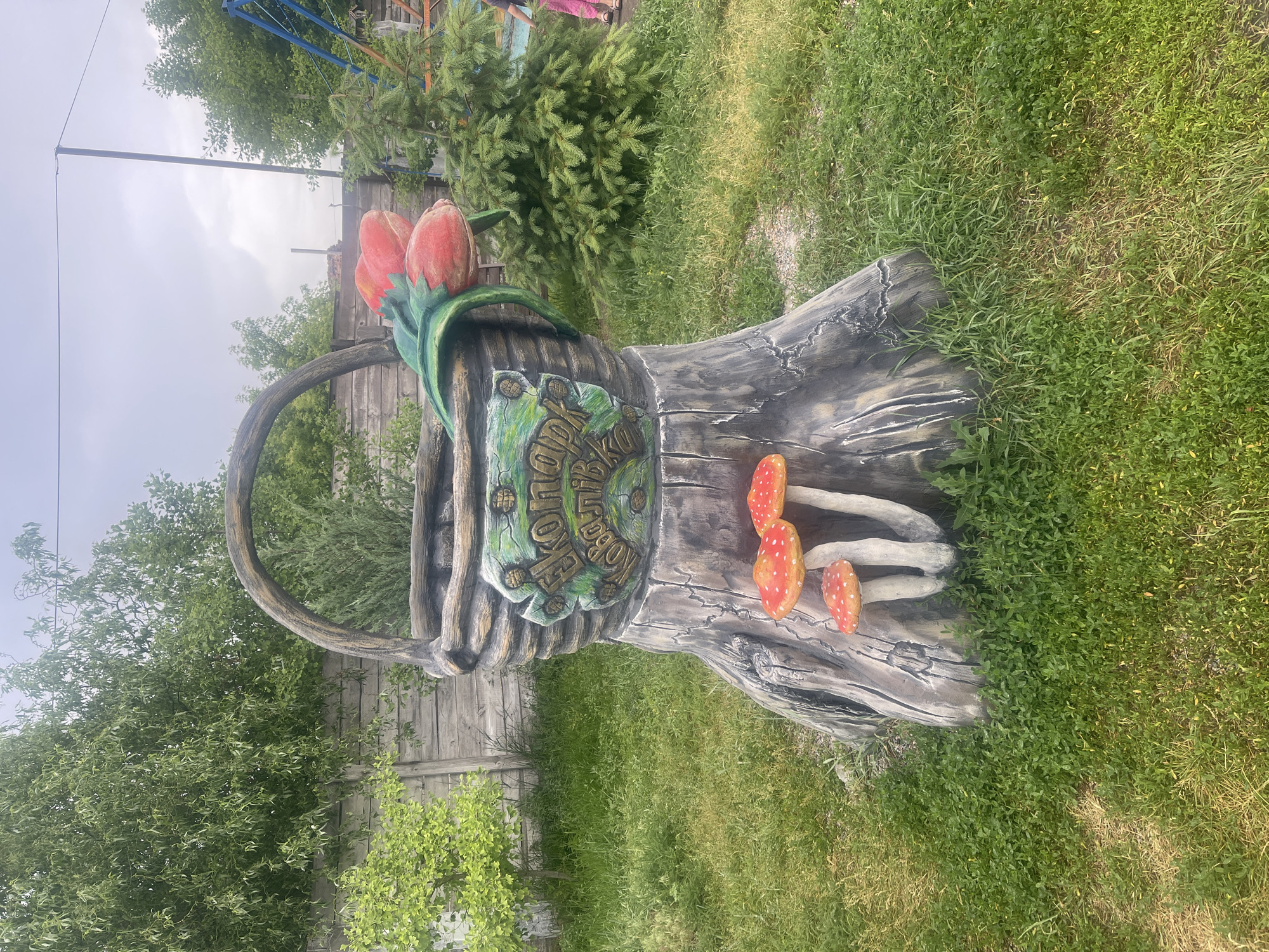 A decorative sculpture resembling a tree stump with a woven basket on top sits in a grassy yard. The basket is adorned with artificial red tulips and a green sign with "Екопарк Ковалівка" inscribed in yellow letters. Several decorative red and white mushrooms are placed at the base of the stump. In the background, a wooden fence, trees, and a swing set are visible. A portion of a person wearing pink pants can be seen on the right side of the image. The sky is overcast.