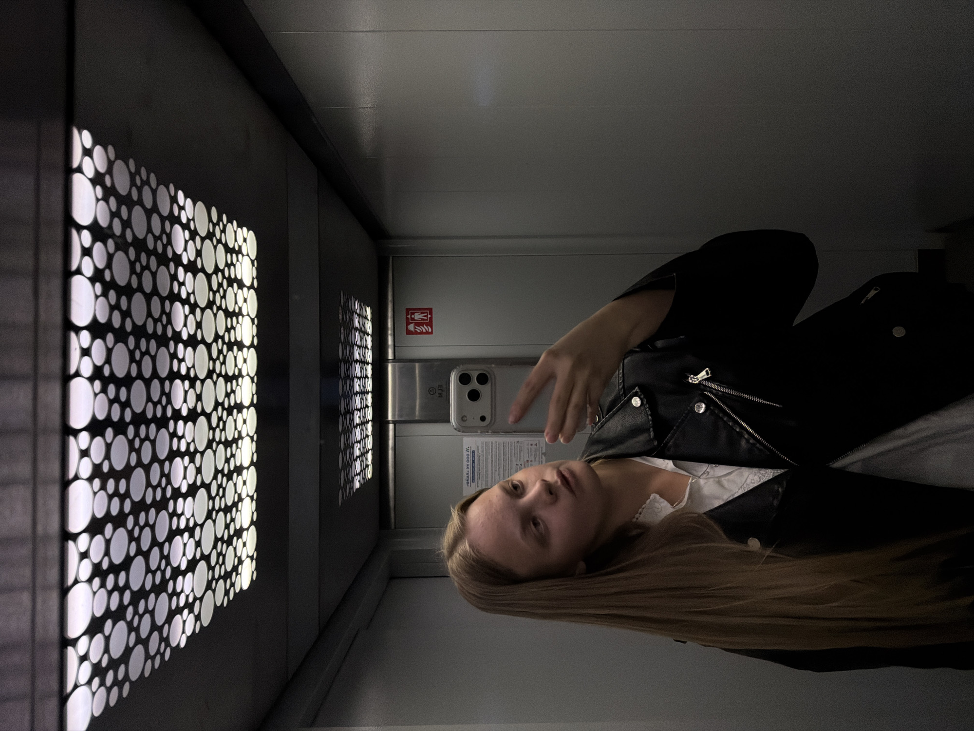 A young woman is depicted in an indoor, dimly lit environment, likely taking a selfie or recording herself with a smartphone held in her right hand. She has long, straight blonde or light brown hair and a neutral to slightly thoughtful expression. She is wearing a dark, possibly leather, jacket over a white top, parts of which show lace detailing.
The setting appears to be a modern corridor or hallway, characterized by dark walls and a light grey ceiling. A prominent feature on the left wall is a decorative, illuminated panel consisting of numerous white circles of varying sizes against a dark background, casting a glow. Another similar panel is partially visible further down the corridor on the right.
On the wall behind the woman, above her shoulder, a red pictogram is visible, resembling an emergency exit or fire safety sign (a figure running, possibly next to an arrow or fire symbol). Below this symbol, on a metallic surface, some text is partially visible, including what appears to be a technical code or serial numbers: "000 К. 37/10" and "77 000 М. 379/8". The smartphone she holds has a light-colored case with three visible camera lenses.
The overall atmosphere is dark, with artificial light sources illuminating the decorative panel and the woman's face from the phone screen. The event takes place indoors, with no discernible time of day or weather elements. The location context provided is Izhevsk, Russia.