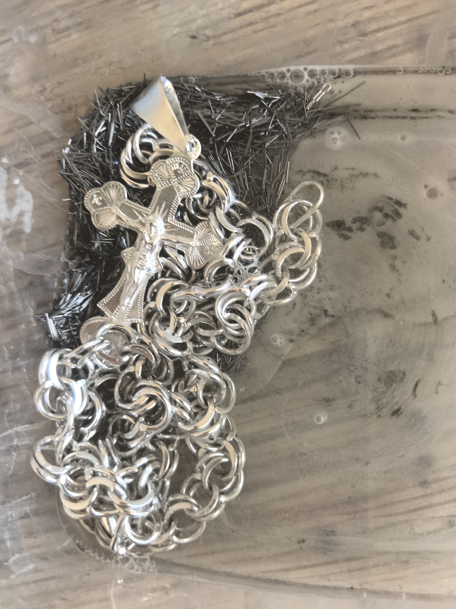 The image shows a silver-colored crucifix pendant with a chain, lying on a pile of small metal rods, possibly nails, submerged in soapy water. The scene is captured from a top-down perspective. The crucifix is detailed, featuring the figure of Jesus. The chain is thick and made of interconnected oval links. The metal rods are numerous and densely packed, appearing dark in contrast to the shiny silver pendant and chain. The water is cloudy and slightly agitated, with some bubbles visible. The background appears to be a wooden surface, likely a table, partially visible beneath the water and objects. There is no indication of the city of Amursk, Russia, within the image.