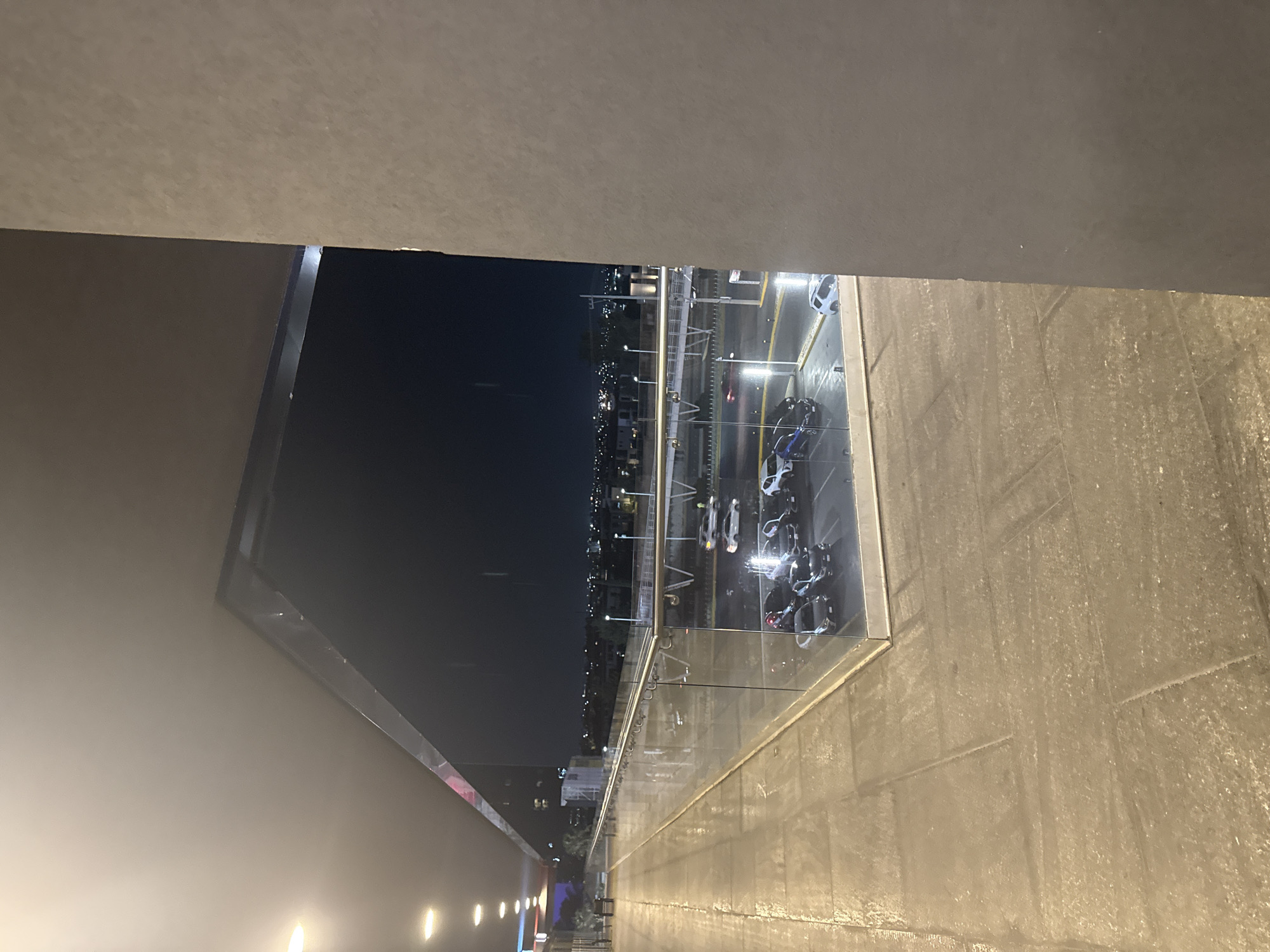 The image captures an elevated, outdoor perspective at night in San Bernardino Tlaxcalancingo, Mexico, overlooking an urban environment. The foreground consists of a tiled pedestrian walkway, appearing wet and reflective, illuminated by recessed overhead lighting. A modern barrier of glass and metal railing runs along the right edge of the walkway. Beyond this railing, a parking lot is visible below, containing approximately eight to ten vehicles, predominantly sedans and SUVs of various dark colors, reflecting ambient light. The asphalt of the parking lot also appears wet. Further into the midground, a multi-lane road with visible streetlights and a pedestrian overpass are present. The background reveals a dark night sky above a sprawling cityscape, marked by numerous distant lights from buildings and streets. To the far right, a vertical section of a textured, light-brown building wall occupies a significant portion of the frame. No individuals are visible in the scene. The overall impression is an urban night setting, possibly after rainfall.