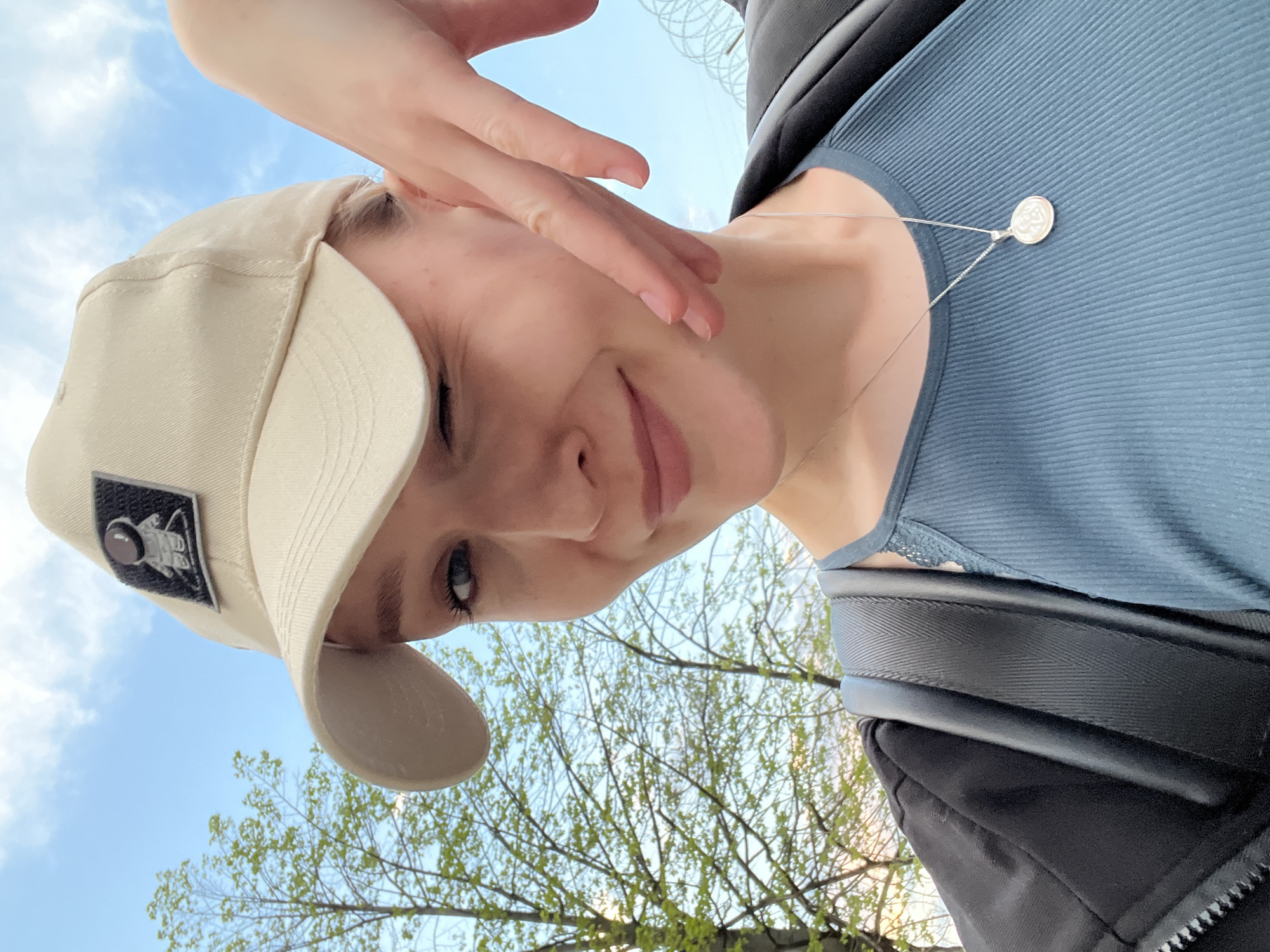 A woman with fair skin wears a beige baseball cap with a black patch featuring a white astronaut emblem. She is wearing a light blue ribbed top and a silver necklace with a circular pendant. Her hand touches her face. The background includes a blue sky with white clouds, and trees with green leaves. The image appears to be taken outdoors. The city is not clearly discernible from the image and cannot be definitively determined as Khimki, Russia.
