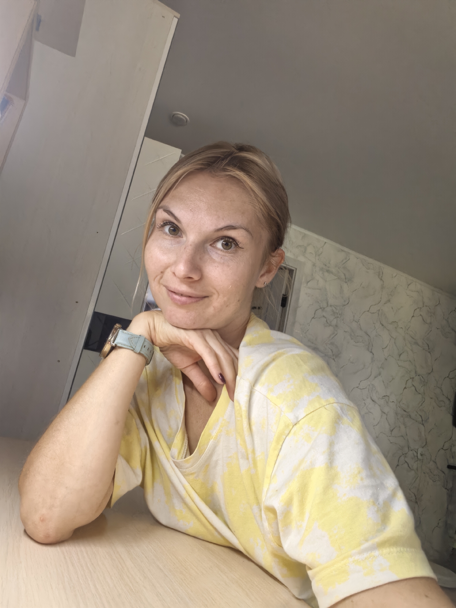 A medium shot from a slightly low angle captures a young Caucasian woman indoors, seated at a light-colored wooden table. Her blonde hair is pulled back loosely, and she has a fair complexion with freckles. She is wearing a yellow and white tie-dye t-shirt. Her right arm is resting on the table, with her hand supporting her chin. A light blue watch is visible on her left wrist. The background consists of a light gray wall with a marbled pattern. To the left, a portion of a light-colored cabinet or closet is visible. The overall lighting suggests an indoor setting with natural light coming from the left. The image does not provide any specific visual cues to indicate a location within Sterlitamak, Russia.