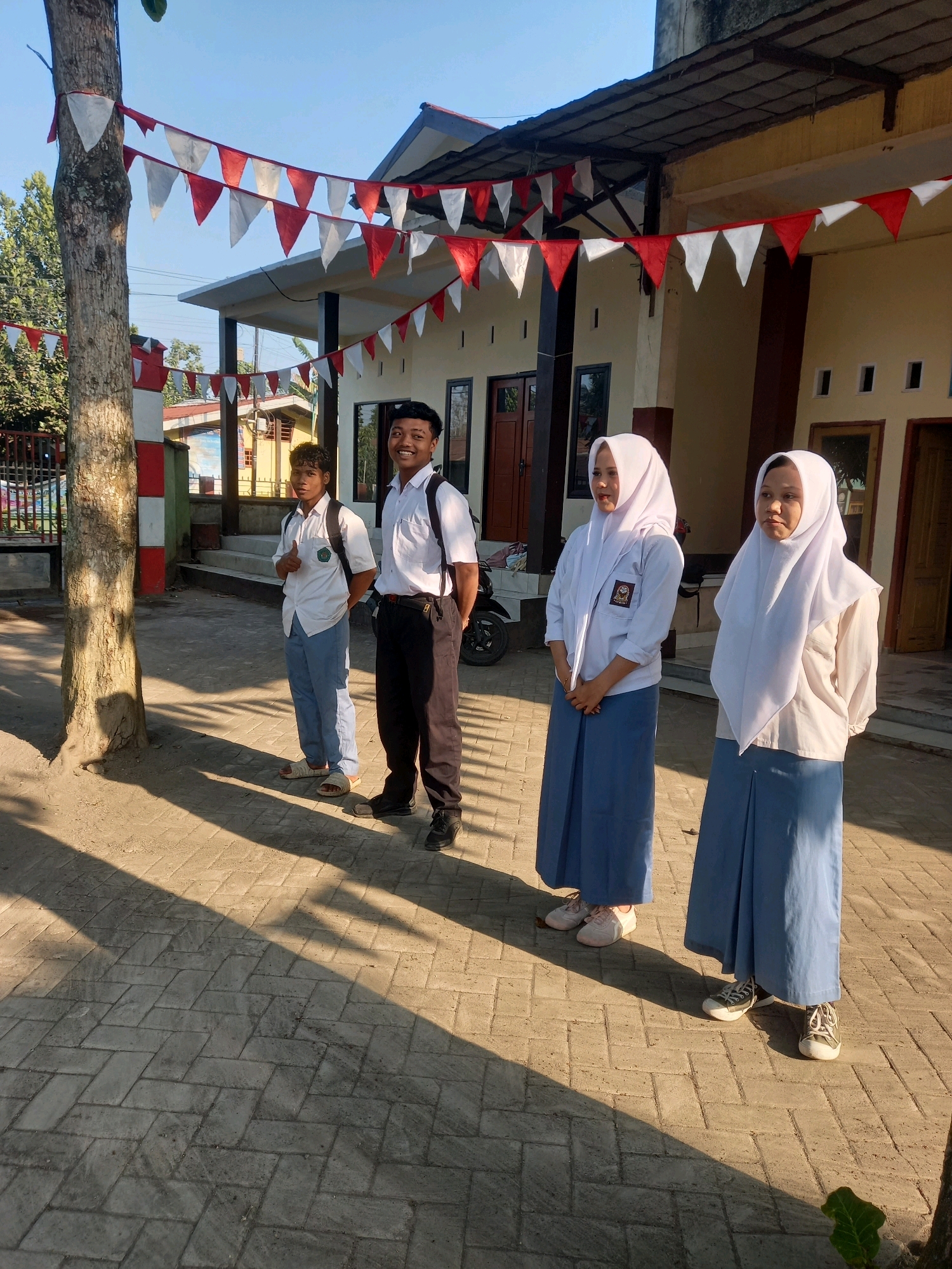 In the city of Perampuan, Indonesia, under a clear blue sky, four students are standing in front of a building adorned with red and white triangular flags. Two male students, dressed in white shirts and dark pants, stand on the left. One is wearing light blue trousers, a white shirt, and a backpack, giving a thumbs-up. The other wears dark pants, a white shirt, and a backpack. To the right, two female students are dressed in white hijabs and long blue skirts, with white shirts. The building appears to be a school or similar institution, with a dark-tiled roof and cream-colored walls. The ground is paved with light-colored rectangular bricks, casting distinct shadows from the sunlight. A large tree trunk with rough bark is visible on the far left of the frame, also decorated with red and white flags. A motorcycle is parked to the right of the male students. The scene suggests a casual outdoor gathering or perhaps the start of a school day.