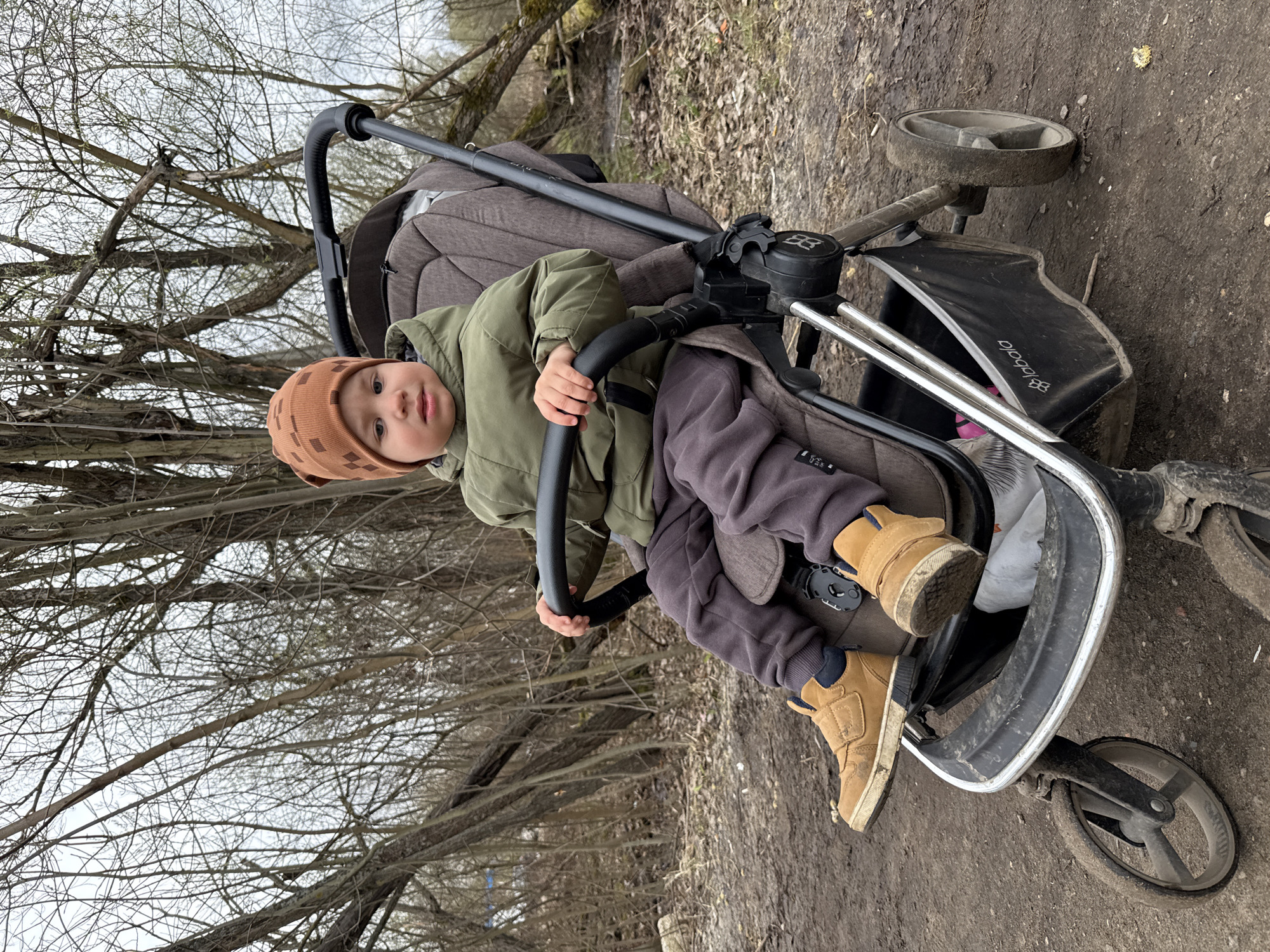 A young child wearing a brown patterned hat, a green jacket, gray sweatpants, and brown shoes is sitting in a stroller. The stroller is on a dirt path in what appears to be a wooded area with bare trees. The ground is muddy, and the sky is overcast. The stroller's brand is "Babystyle". The location is in Rusanovo, Russia.