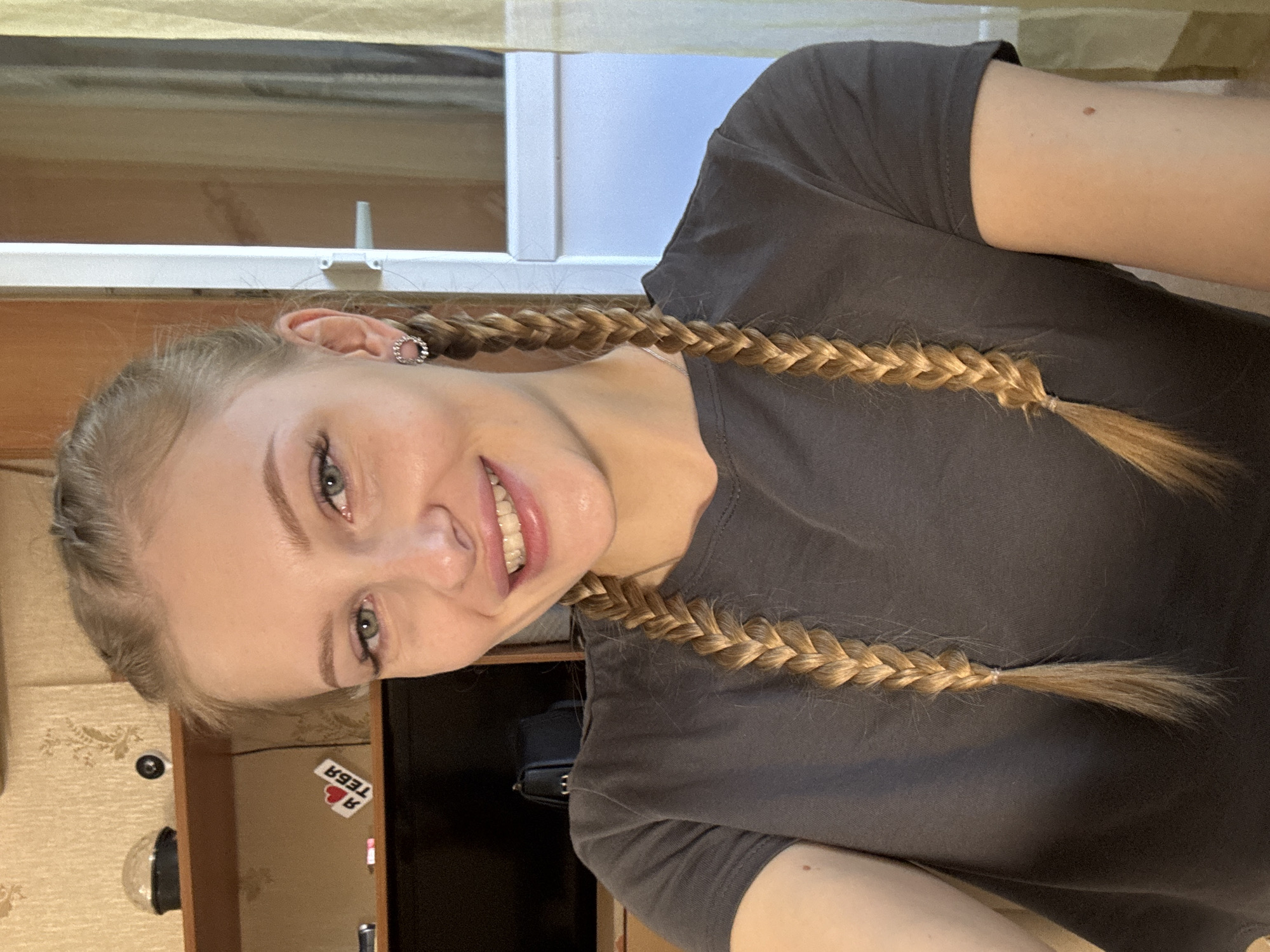A young woman with blonde hair styled in two braids is shown in a close-up, chest-up shot. She is wearing a dark grey t-shirt and a circular earring in her visible ear. She is smiling and looking directly at the camera. Behind her, a shelf is visible with a decorative object and a sticker that says "Я тебя" which translates to "I love you" in Russian. The background suggests an indoor setting, likely a room, with a glimpse of a doorway or window with curtains. The overall lighting is bright, indicating a well-lit indoor space. There is no identifiable information within the image that places this scene specifically in Yekaterinburg, Russia.