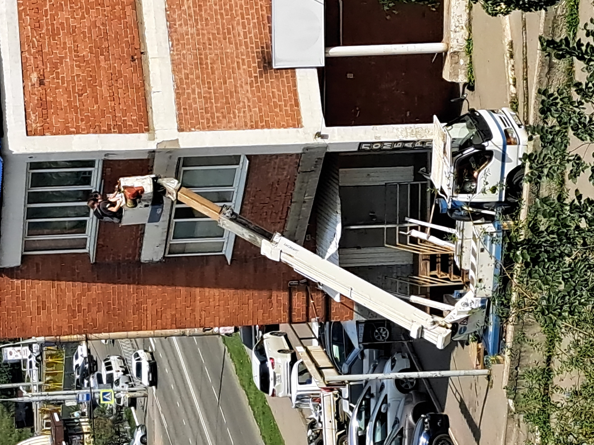 This image captures an urban scene in Irkutsk, Russia, with a focus on a building undergoing external work. A white lift truck, with its mechanical arm extended, is positioned next to a multi-story brick building. At the top of the extended arm, a white bucket holds two individuals, seemingly engaged in tasks on the building's facade. One person is sitting, while the other is standing, both looking downwards. Below them, the building has several windows and a section of exposed brickwork. A covered walkway or awning is visible beneath the working area. In the background, a road with several parked and moving vehicles, including cars and a white van, suggests the location is a public thoroughfare. Trees and vegetation are present in the foreground and along the roadside. The scene appears to be set during daylight hours.