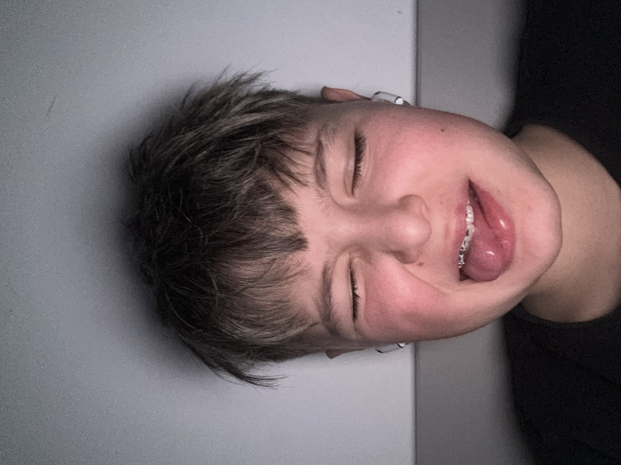 A young person with short, choppy brown hair lies with their head tilted on a gray surface. The person's eyes are closed, and their tongue is sticking out, showing teeth with braces. A small white object, possibly an earbud, is visible near the ear. The person is wearing a dark shirt. It is not possible to determine the location as Sofia, Bulgaria, without additional information from the image.