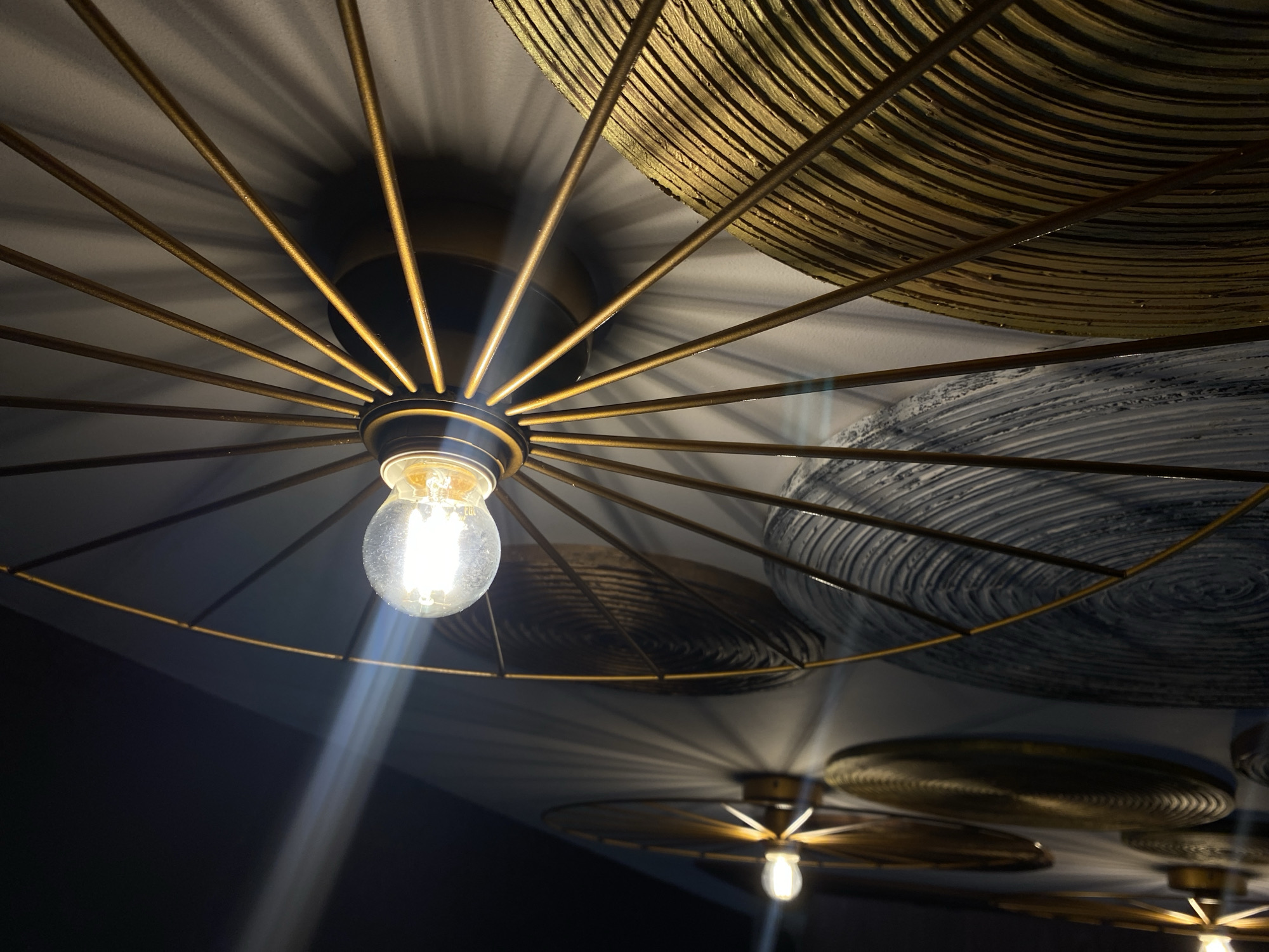 A low-angle interior shot features a light fixture with a visible lightbulb at its center, surrounded by a golden, spoked frame. Shadows cast by the frame are visible on the ceiling. Several decorative, circular objects with textured surfaces are also affixed to the ceiling, and their shadows are apparent on the surface as well. Another, similar light fixture with a lightbulb is visible in the background. The location is not discernible.