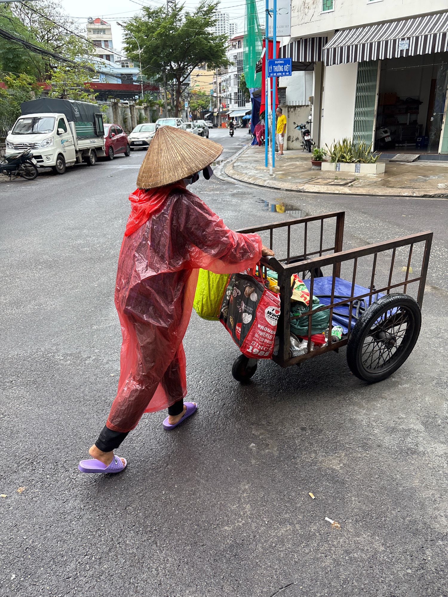 A person wearing a red disposable raincoat and a conical hat is pushing a metal cart filled with various items along a wet asphalt street in Nha Trang, Vietnam. The cart has two wheels, one of which is a spoked bicycle wheel. The person is also carrying a bright green bag and a black bag. In the background, there are buildings, vehicles including a white truck and a red car, and trees. The street appears to be a commercial area with storefronts and awnings. There are two cigarette butts on the ground in the lower right portion of the image. The scene suggests a rainy or recently rained-on day.
