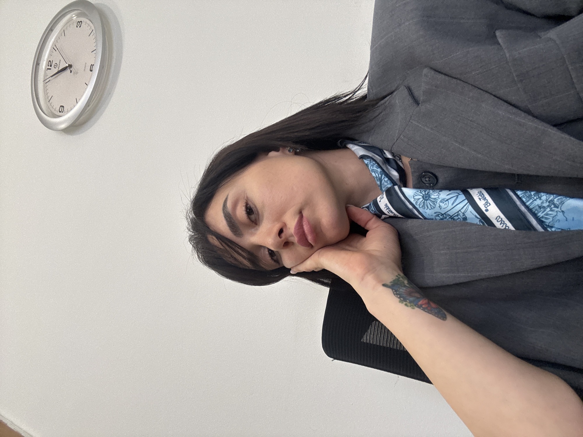 A medium shot of a woman posing with her head resting on her hand. She is wearing a gray pinstripe blazer and a blue patterned scarf. Her dark hair is neatly styled. A clock hangs on the white wall behind her, showing the time as approximately 12:12. A butterfly tattoo is visible on her arm. The scene is brightly lit. There is no information to specify the location as Tbilisi, Georgia.