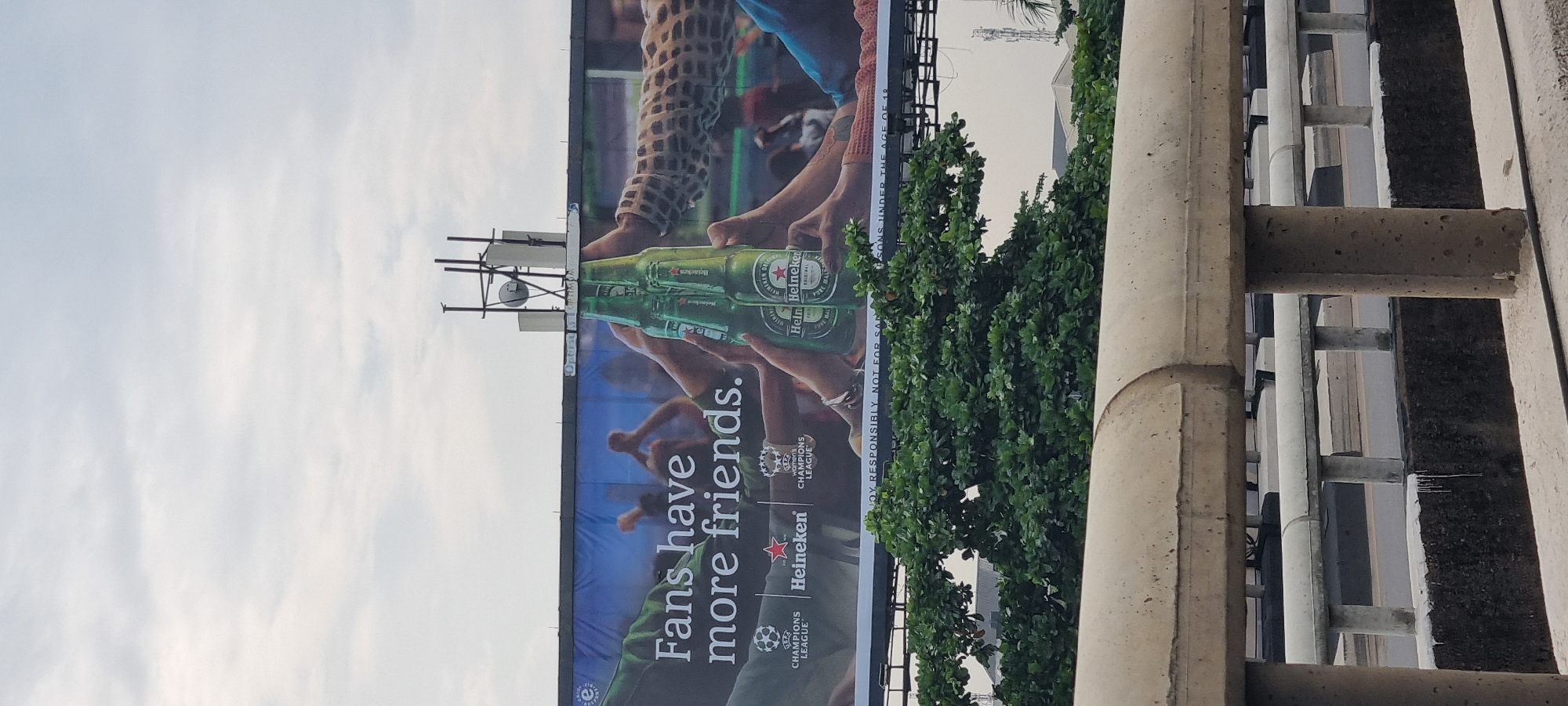 The image depicts a billboard on the outskirts of Alagbede, Nigeria, featuring a Heineken beer advertisement. The billboard's text reads "Fans have more friends" and prominently displays Heineken branding along with UEFA Champions League logos, indicating a partnership. Several hands are shown clinking green Heineken bottles together in a celebratory gesture, with some hands displaying patterned sleeves. The background of the billboard shows blurred figures, likely of fans, in a celebratory mood. Below the main advertisement, a disclaimer states, "ENJOY RESPONSIBLY. NOT FOR SALE TO PERSONS UNDER THE AGE OF 18."
The billboard is erected on a metallic frame and is partially obscured by lush green foliage from trees in the foreground. In the background, a clear sky with light clouds suggests daytime. A concrete barrier with a white railing runs across the lower part of the image, implying the perspective is from a bridge or elevated roadway overlooking the scene. The overall environment appears to be an urban or semi-urban setting, characterized by the advertising structure, the foliage, and the presence of the roadway.