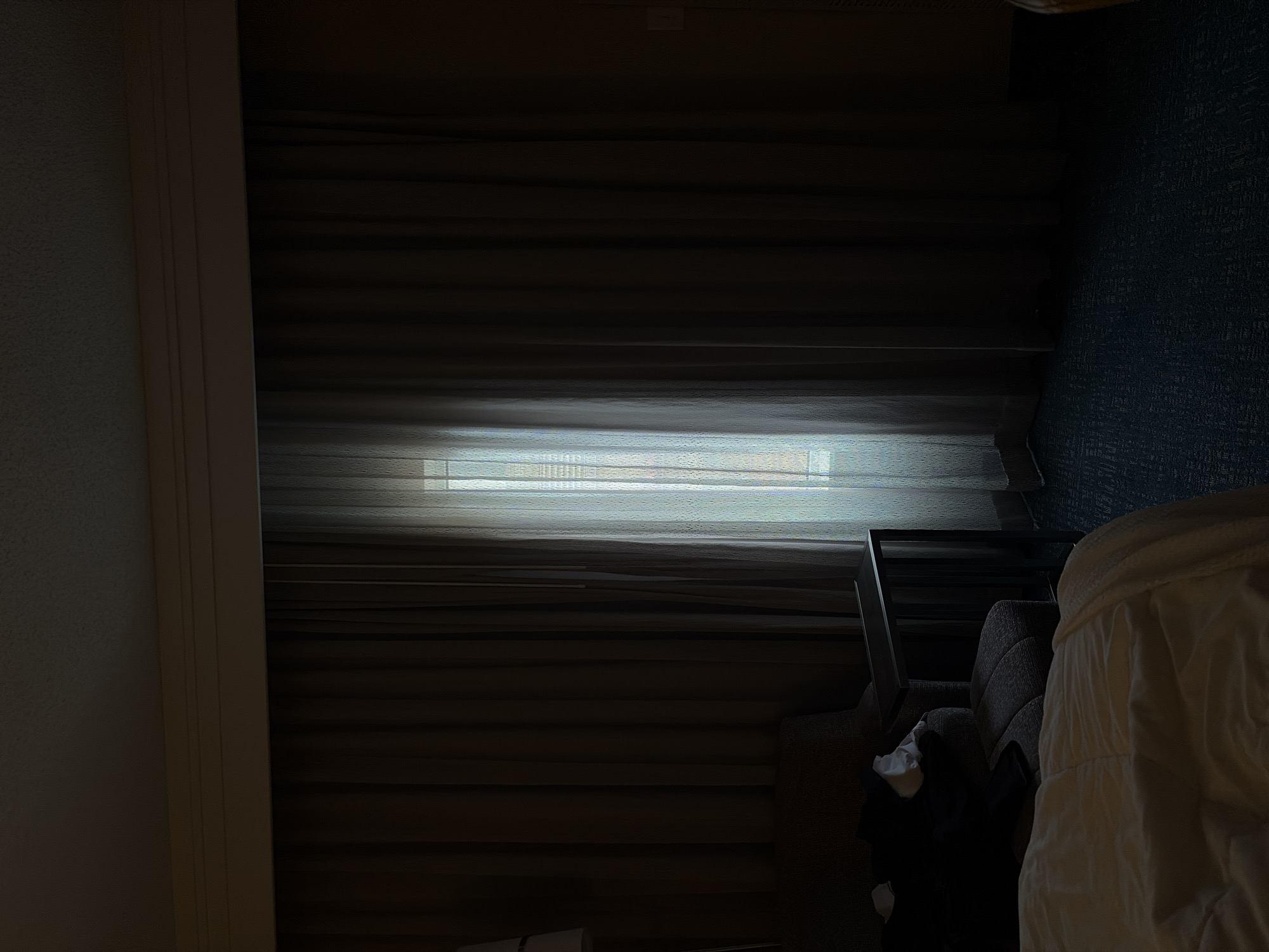 The image shows the interior of a room in San Antonio, United States of America. The room is dark, with light filtering through closed blinds on a window. A chair with a metal frame is partially visible on the right side of the frame, next to what appears to be a bed with white linens. A black garment is crumpled at the base of the chair. The walls have a textured surface.