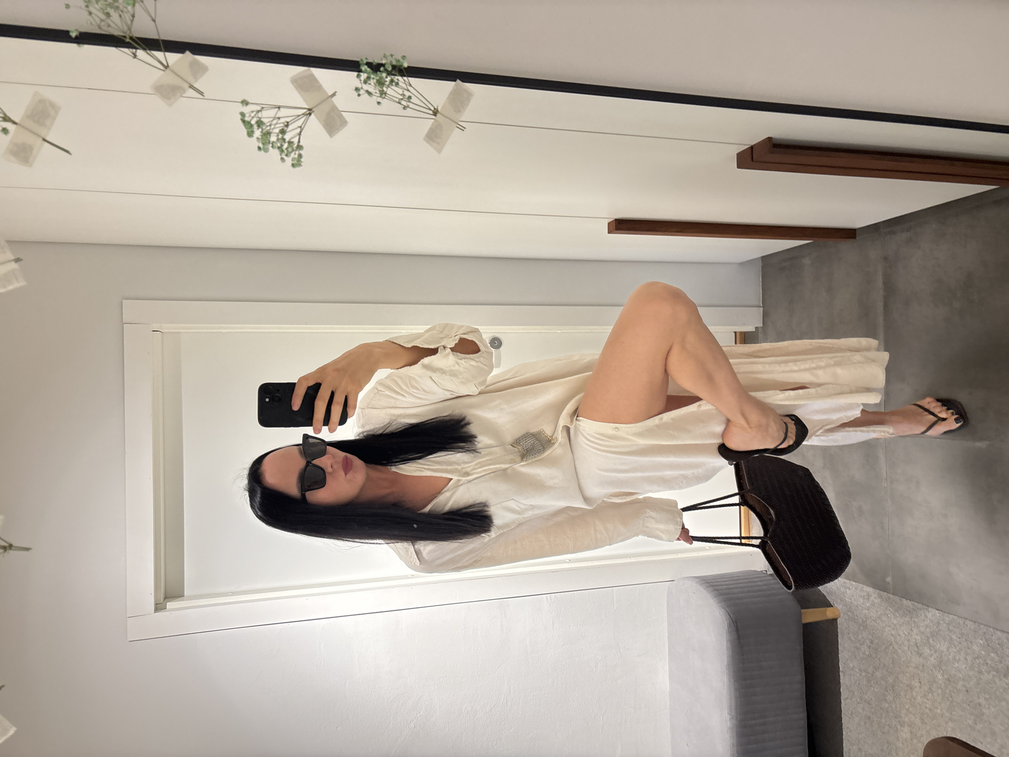 A woman with long, dark hair and sunglasses is posing for a mirror selfie. She is wearing a white, button-down dress, flip-flops, and holding a black purse. She is holding up her phone in her right hand to take the photo. The room appears to be a bedroom or a hotel room with a grey couch and a white door. The wall is white and a mirror is decorated with baby's breath flowers. The location is unclear, but the city of Chaikovsky, Russia, is provided.