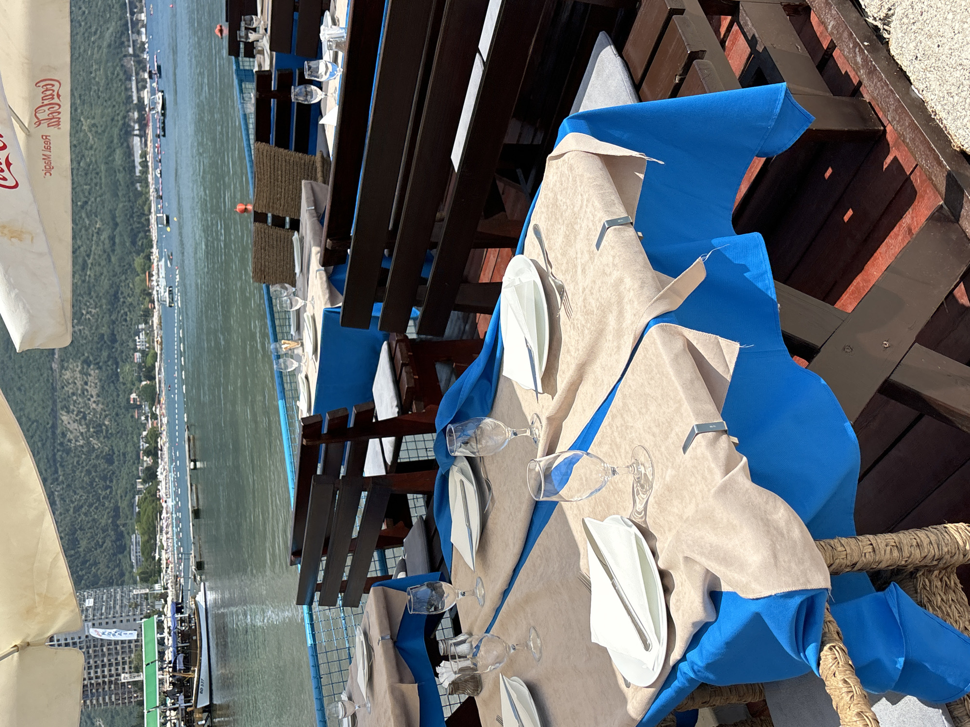 This image depicts an outdoor seating area of a restaurant or cafe, likely situated in Budva, Montenegro, given the coastal scenery. The foreground features several tables set for dining, covered with blue tablecloths topped with beige runners. Each place setting includes a white plate with a folded white napkin and silverware, accompanied by clear glassware. Wooden benches with grey cushions are positioned behind the tables.
In the mid-ground, the Adriatic Sea stretches out, with a clear blue sky above. Several boats are visible on the water, and a promenade or walkway lines the shore, with buildings and beach umbrellas indicating a bustling tourist area. The background reveals a mountainous landscape behind the coastal town. The overall atmosphere suggests a sunny day in a popular seaside resort.