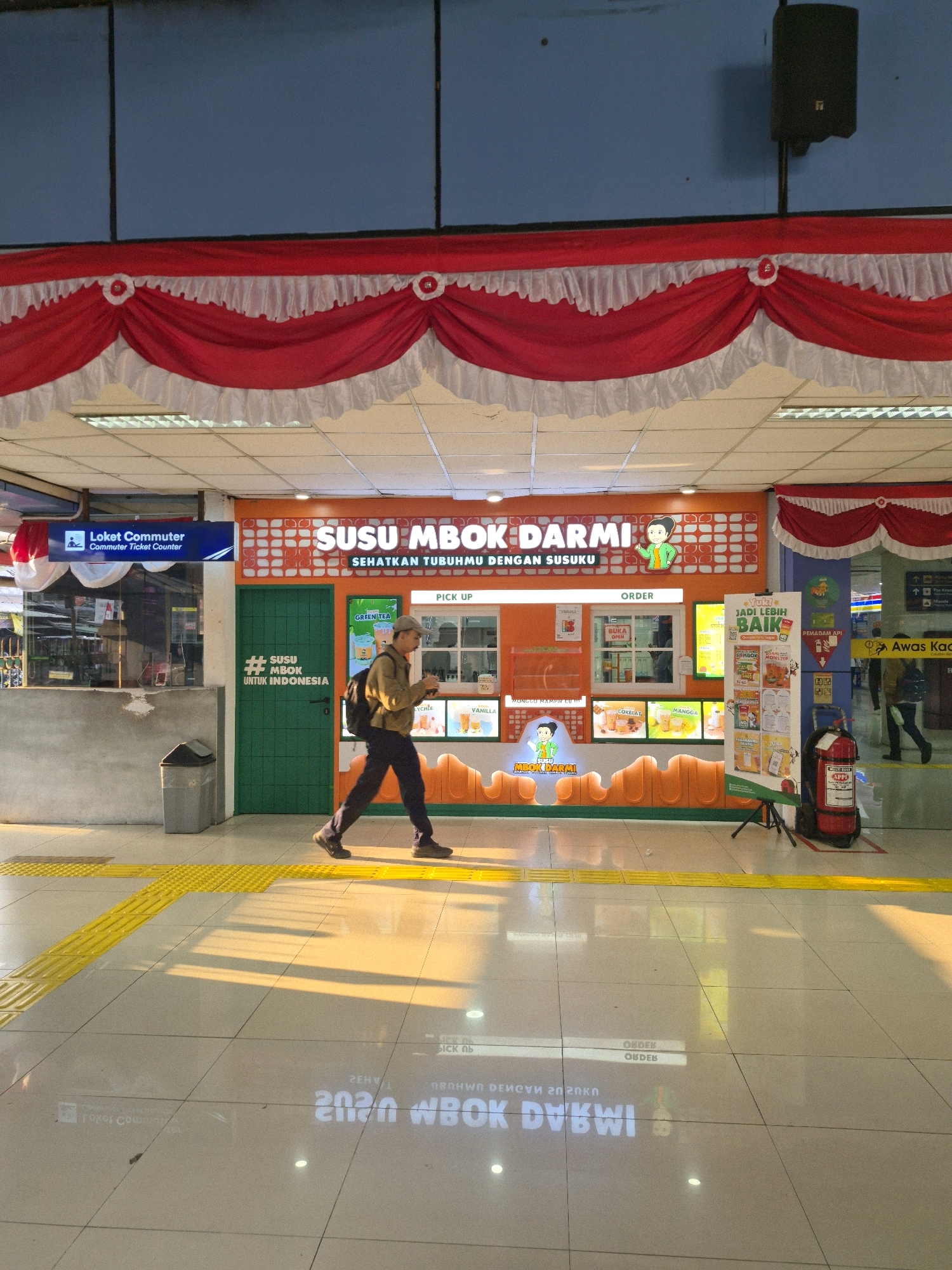 The image shows a man walking in front of a brightly colored food stall called "SUSU MBOK DARMI" located in Central Jakarta, Indonesia. The stall is decorated with orange and white tiles and has various drinks displayed. The man is wearing a light-colored jacket, dark pants, and a backpack, and he appears to be looking at his phone. The floor is tiled, and there are yellow tactile paving strips to guide visually impaired pedestrians. Red and white draped fabric banners are hung above the stall, suggesting a festive or nationalistic theme. A fire extinguisher is visible to the right of the stall. The lighting indicates it is daytime, with strong sunlight casting shadows on the floor.
