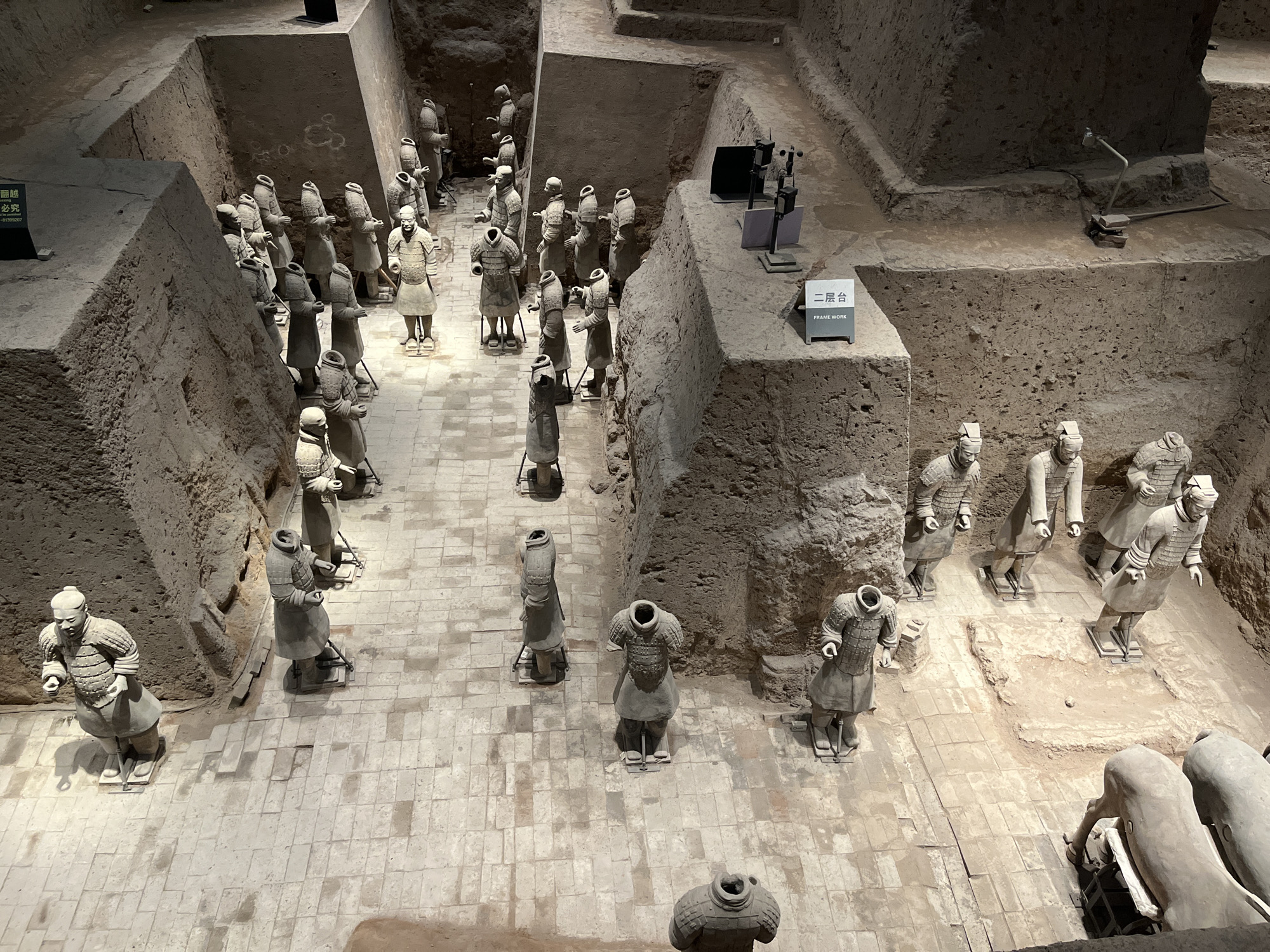 The image depicts the Terracotta Army, a collection of terracotta sculptures depicting the armies of Qin Shi Huang, the first Emperor of China. The scene is an archaeological excavation, with the life-sized figures arranged in rows and columns, as if in formation. Many of the figures are soldiers in armor, some with helmets, while others appear to be generals or officers, distinguished by their attire. Some figures are headless, indicating the passage of time and the effects of excavation and preservation. The figures are made of terracotta, with visible details of their clothing, armor, and facial features. The environment is characterized by excavated earth walls and a tiled floor. There are informational signs in Chinese and English visible in the image, one of which reads "FRAME WORK." The lighting suggests an indoor or covered exhibition space, illuminating the figures and the surrounding excavation pit. The overall impression is one of a vast, ancient army frozen in time.