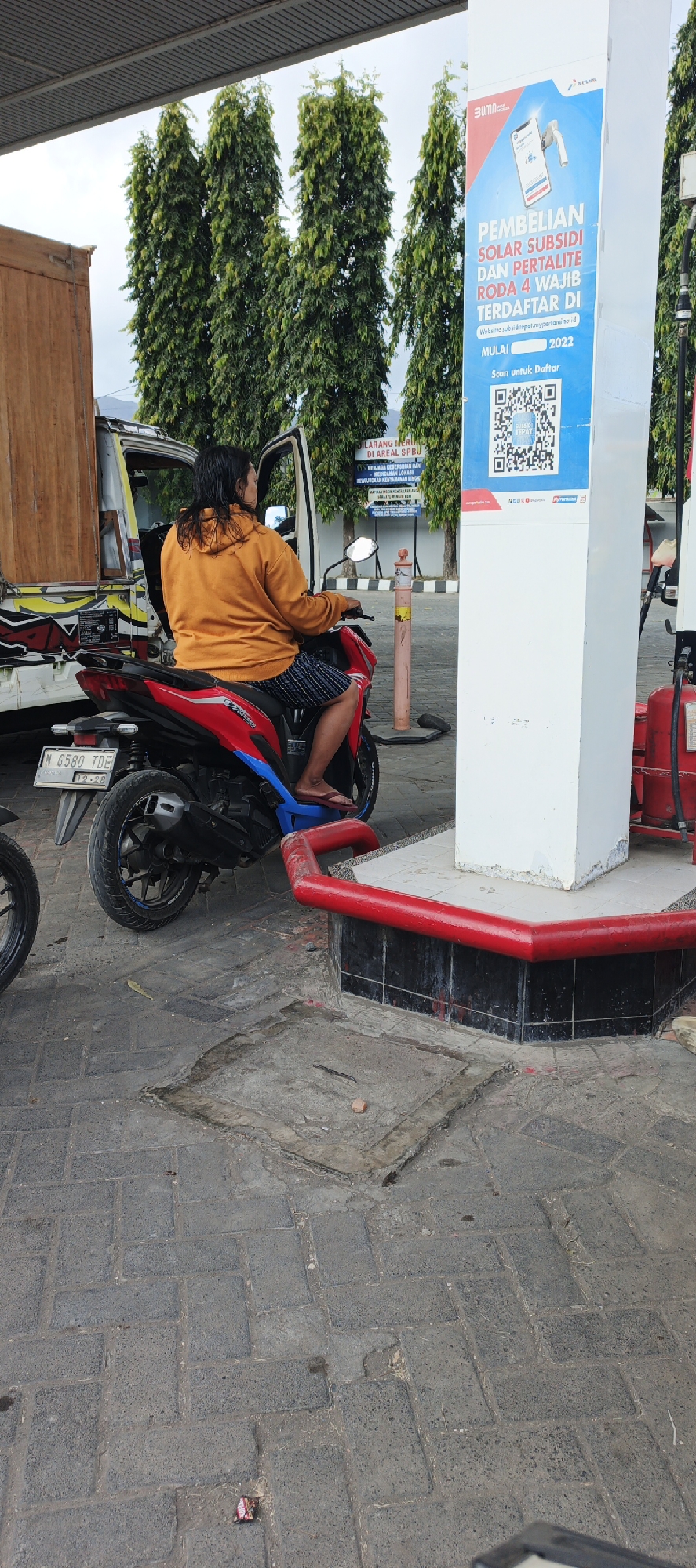 The image depicts a scene at a gas station in Grenden, Indonesia. A woman is sitting on a red motorcycle, facing away from the camera, with the motorcycle's engine cover open. She is wearing an orange hoodie and checkered shorts. Behind her, a white truck with a wooden cargo bed is parked. A prominent blue and white sign on a white pillar, likely a gas pump, displays information in Indonesian about purchasing subsidized solar and Pertalite for four-wheeled vehicles, requiring registration through a website or QR code scan. The ground is paved with interlocking gray bricks, and there are trees in the background. A red guardrail is visible around the base of the white pillar. The overall impression is a typical refueling stop.