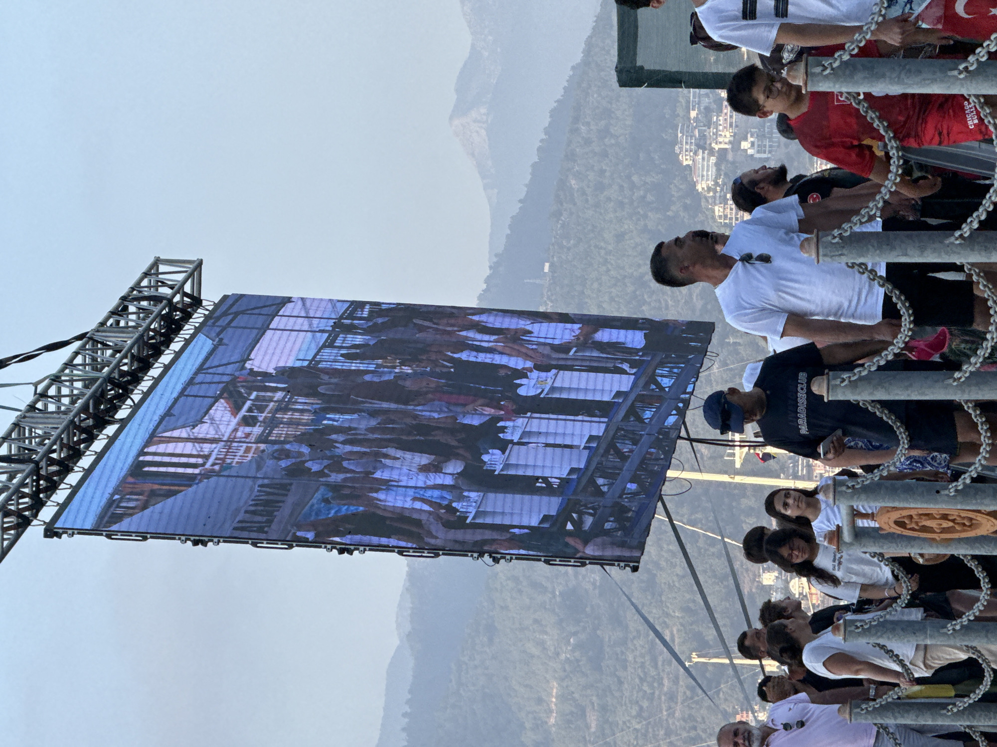 A large outdoor LED screen, displaying an image of a crowd and the word "ALANYA", is suspended from a metal truss structure. The screen is tilted and appears to be part of an event in Alanya, Turkey, given the visible text. In the foreground, a group of people are standing behind a chain barrier. Among them, a man in a white t-shirt and black shorts is looking towards the screen. To his right, a boy in a red t-shirt and shorts is also looking downwards. Behind them, other individuals are visible, some interacting with each other. The background features rolling, tree-covered hills with buildings scattered on their slopes, suggesting an urban or semi-urban setting. The sky is overcast, and the overall lighting indicates it is daytime.