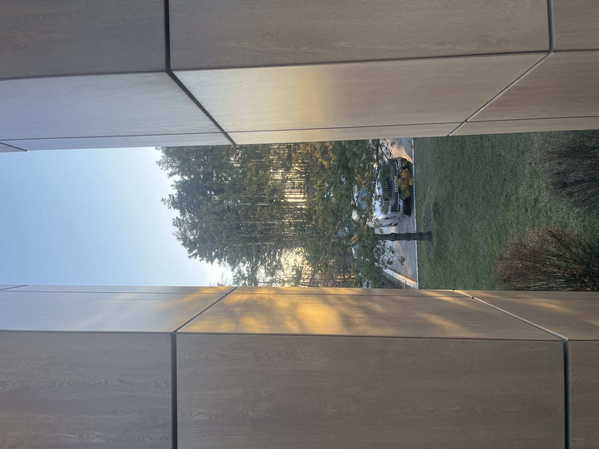 A view from within a building towards an outdoor scene in Zhavoronki, Russia. The perspective is framed by the building's interior elements, possibly walls or dividers composed of large, rectangular wooden or wood-like panels. These panels are primarily a neutral, brownish-gray color. The outdoor scene reveals a glimpse of trees, grass, and parked vehicles. The trees appear to be slender and the sunlight is visible through the trees. There are cars parked along the side. A patch of green lawn is visible. The sky is bright and clear blue in the upper portion. The overall image conveys a sense of looking outward from an interior space.