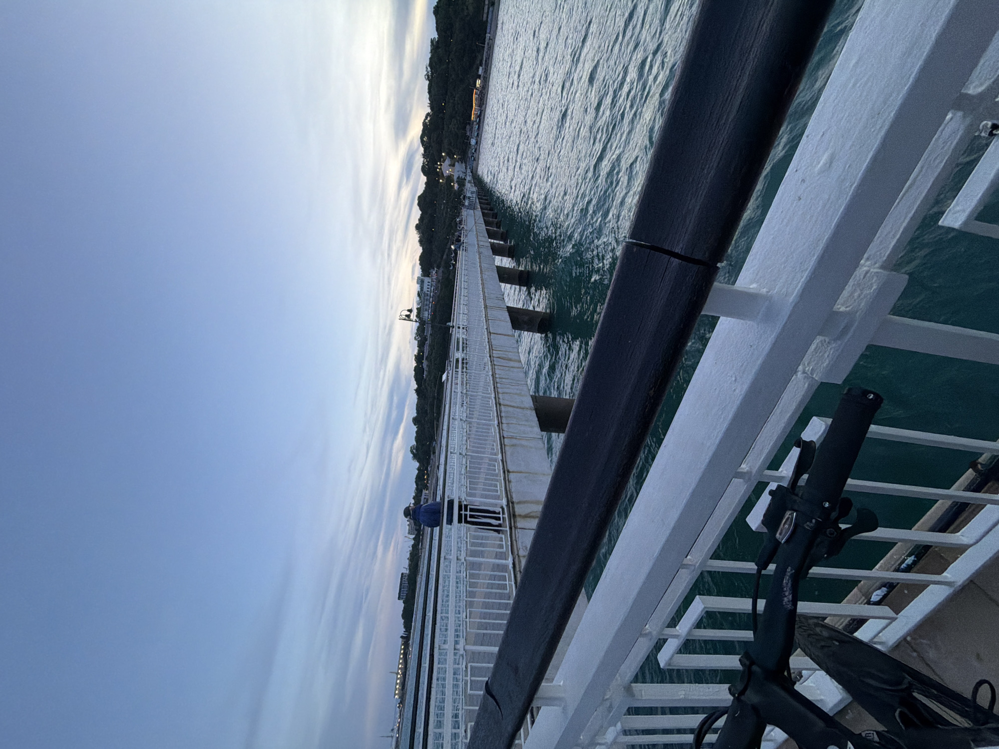 The image depicts a pier extending over the sea. The pier has white railings and a dark handrail in the foreground. A person in a blue jacket and dark pants is standing on the pier, facing away from the camera, looking towards the sea. The sea is a deep turquoise color, with visible ripples. In the distance, a tree-covered coastline is visible, with buildings and lights indicating a town or city. The sky is a pale blue with scattered wispy clouds. In the immediate foreground, the handlebars and front wheel of a bicycle are visible, suggesting the image was taken from a stationary bicycle on the pier. The overall scene is calm and suggests it may be late afternoon or early evening. Based on the provided context of the city of Burgas, Bulgaria, this pier is likely the Burgas Sea Station Pier, a popular promenade and transportation hub.