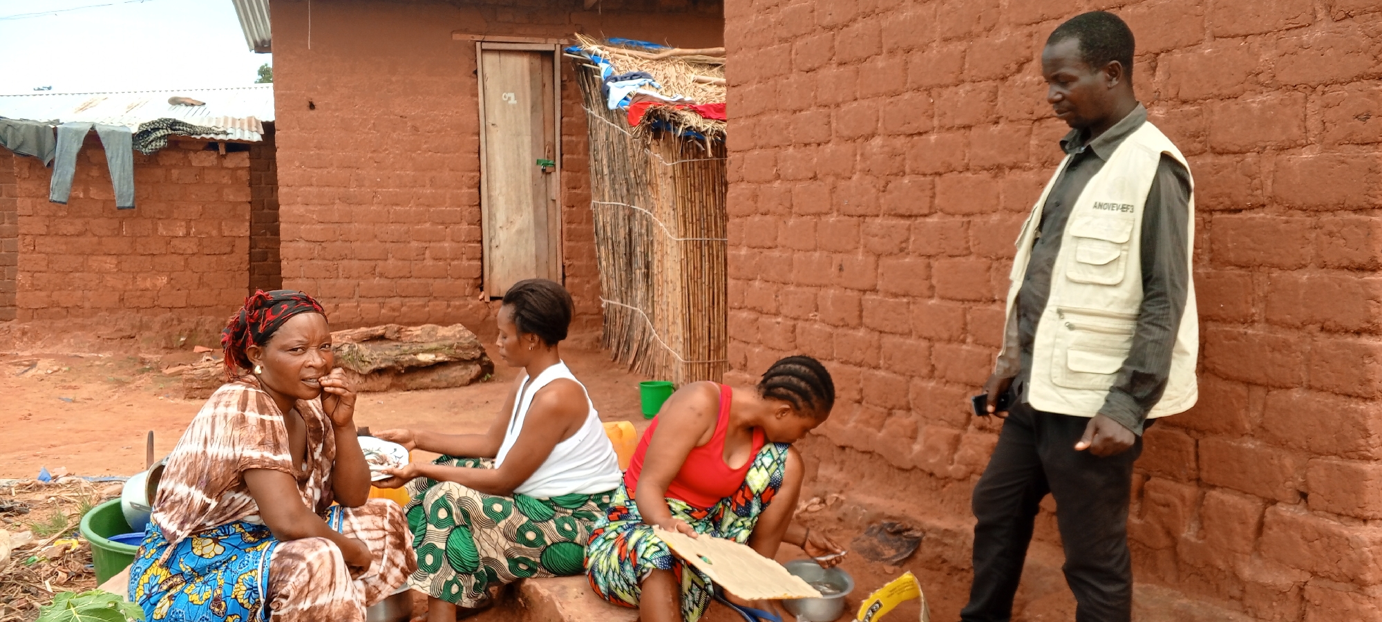 The image shows four individuals outside a brick building in Makungu, Democratic Republic of the Congo. Three women are seated. The woman on the left is wearing a patterned brown and blue dress with a headscarf. She is seated on a green bucket. The woman in the center is wearing a white tank top and a green patterned skirt. The woman on the right is wearing a red tank top and a patterned skirt, and appears to be preparing food. A man in a vest is standing to the right of the women, holding a cell phone. The brick building has a door and a thatched roof extension. Laundry is hanging on a line in the background.