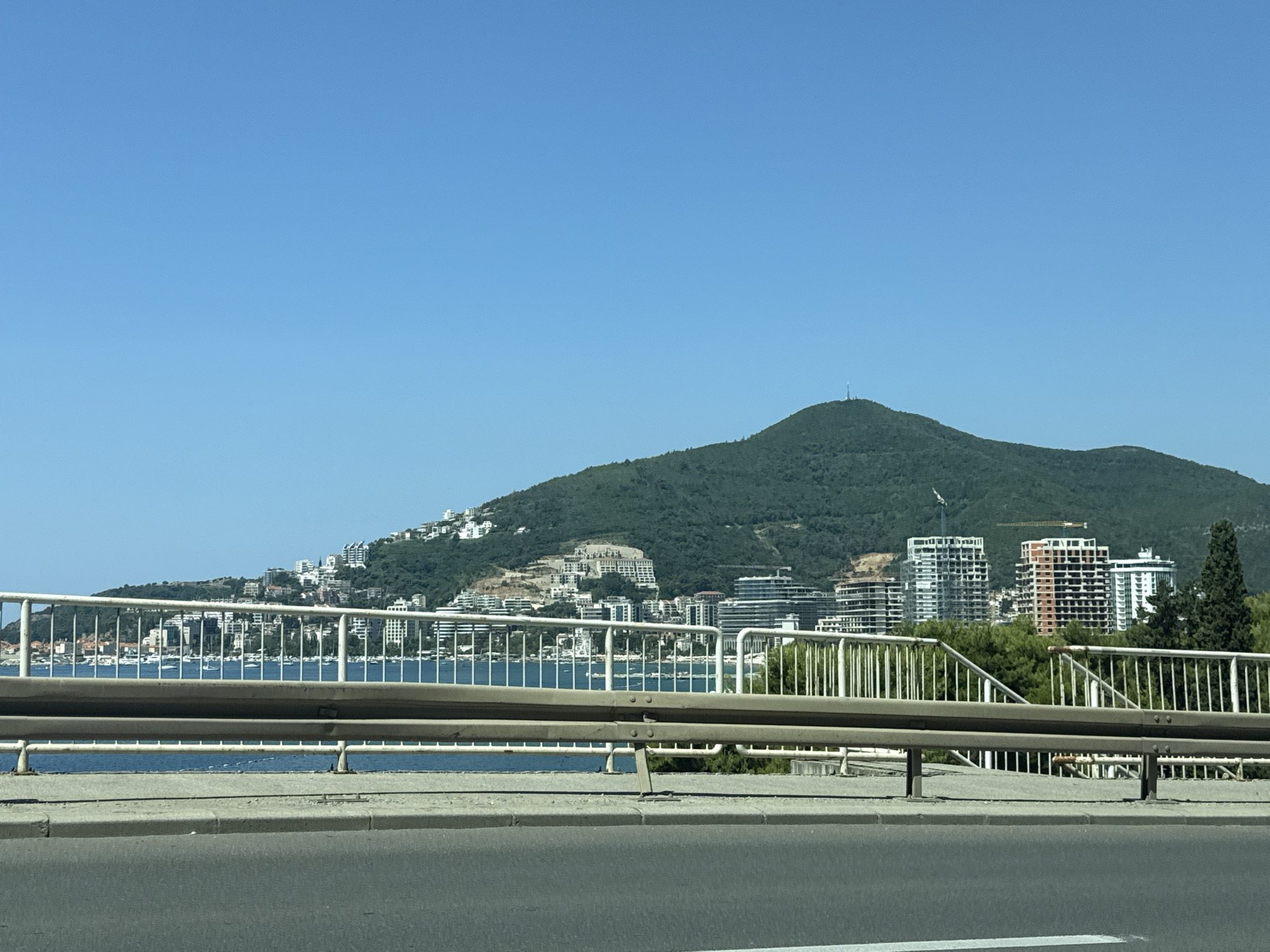 This media file captures a view from a bridge overlooking a coastal city and a forested mountain. The foreground shows a concrete road and a metallic guardrail, suggesting the perspective is from a vehicle in motion. Beyond the bridge, a body of water, likely the sea, is visible, dotted with numerous boats.
The middle ground features a densely built urban landscape cascading down the hillside, characterized by white and light-colored buildings, some appearing to be hotels or apartment complexes. Construction cranes are visible among some of the newer structures, indicating ongoing development. The architecture suggests a Mediterranean or European coastal town.
Dominating the background is a large, verdant mountain, its slopes covered in dense green foliage. The summit of the mountain is crowned with a tall antenna or communication tower. The sky above is a clear, bright blue, indicative of a sunny day. The overall scene evokes a sense of a vacation destination, possibly in the late morning or early afternoon, given the bright light and clear visibility. There is no visible text in the image. The location context provided is Budva, Montenegro, which aligns with the visual cues of a coastal city nestled against a mountainous backdrop.