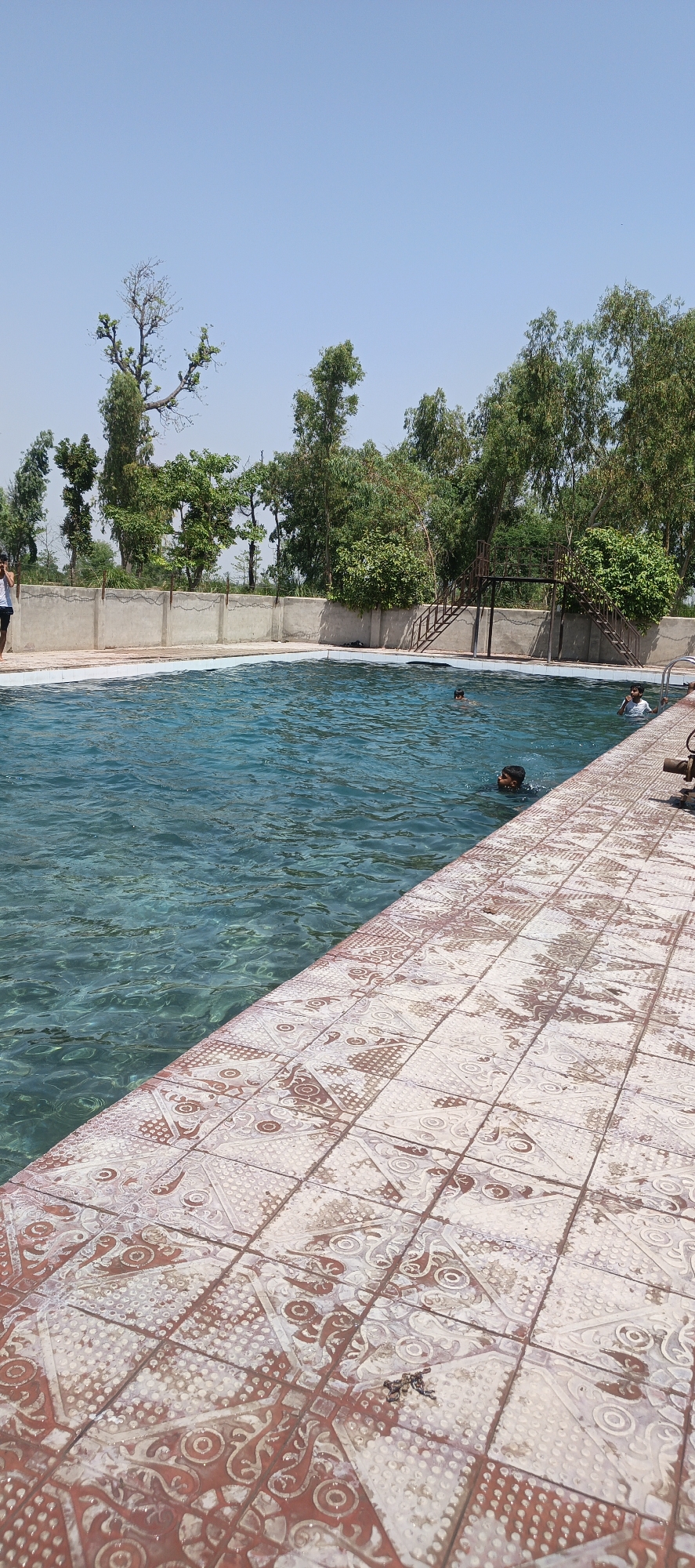 The image shows a swimming pool in Samundri, Pakistan. The pool is rectangular, with clear blue water. There are three people swimming in the pool, one is near the camera and close to the pool edge. The pool is surrounded by a tiled patio and a low concrete wall. Behind the wall, there are several trees. A metal structure with stairs leads to a platform is along the back edge of the pool. The sky is clear and blue.