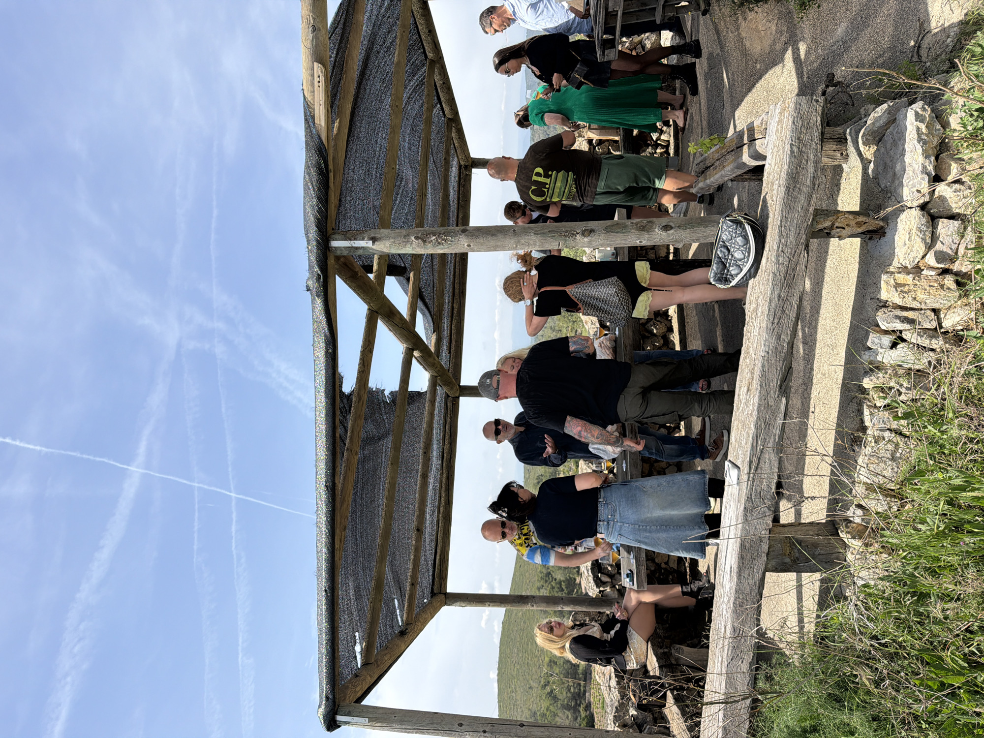 A group of approximately fifteen people are gathered outdoors under a wooden shelter with a dark, mesh-like covering, likely providing shade. The image, set in Bonastre, Spain, shows individuals of various ages, mostly adults, engaged in a social gathering. Some are standing, while others are seated or leaning against wooden structures that form part of the shelter and a viewing platform.
The subjects are dressed casually, with t-shirts, shorts, jeans, and skirts visible. Notable clothing items include a man wearing a black t-shirt with "C.P. COMPANY" printed in yellow, and a woman in a green pleated dress. Several people are holding clear cups, suggesting they are enjoying beverages. Various bags are visible, including a black quilted handbag (possibly Dior) and a grey patterned tote bag. A white personal transport device, resembling a hoverboard, is near the feet of one individual.
The setting appears to be an elevated viewpoint, overlooking a hilly landscape covered in green vegetation, visible in the background. The immediate foreground features rugged stone walls and dry grass, indicative of a natural, somewhat rustic environment. The ground beneath the people is a mix of concrete or packed earth and exposed stone.
The sky is bright blue with several prominent white contrails from aircraft, suggesting clear weather. The strong sunlight casts distinct shadows, indicating it is likely midday or early afternoon. People generally appear relaxed and engaged, either in conversation or observing the view.