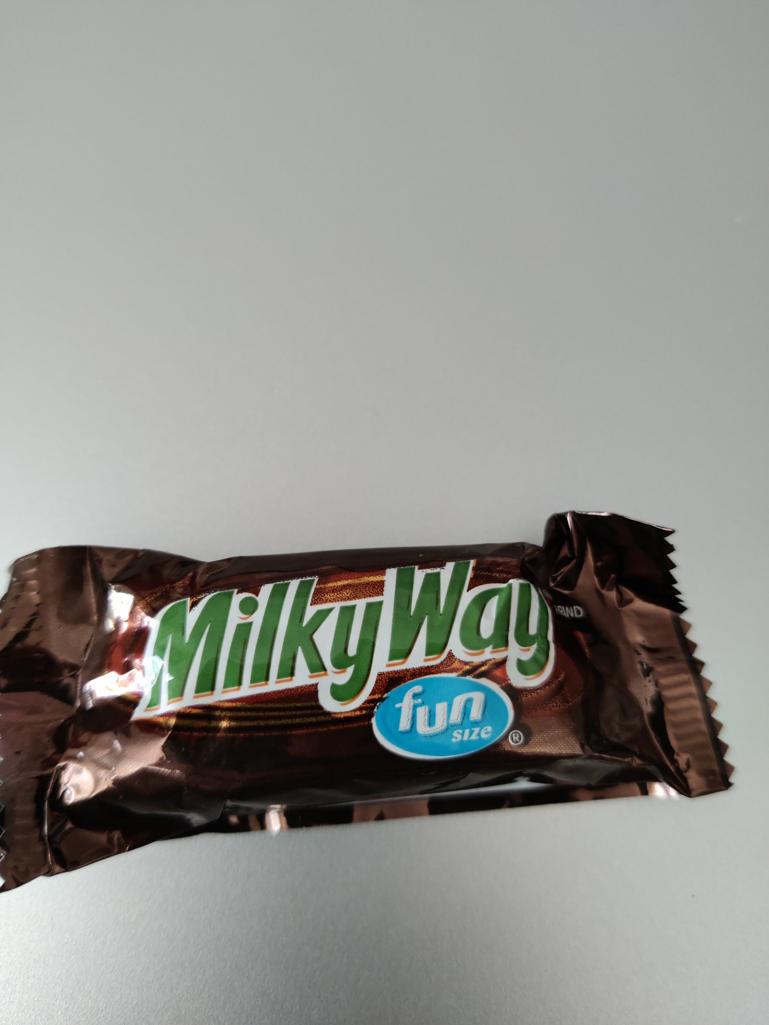 A Milky Way candy bar wrapper is positioned horizontally on a light gray, textured surface, which appears to be a desk or table. The wrapper is made of reflective, dark brown material and is creased and slightly crumpled, indicating it is likely a single-serving fun size wrapper. The prominent logo on the wrapper features the words "Milky Way" in a stylized green font with white outlines. Below "Milky Way" is a small, light blue oval with the word "fun" in white, followed by "size" in smaller white text. A registered trademark symbol is also visible within the blue oval. The background behind the candy bar wrapper is a plain, solid light gray. The image was captured with a neutral overhead lighting setup. The location is identified as Lower Paxton Township, United States of America.
