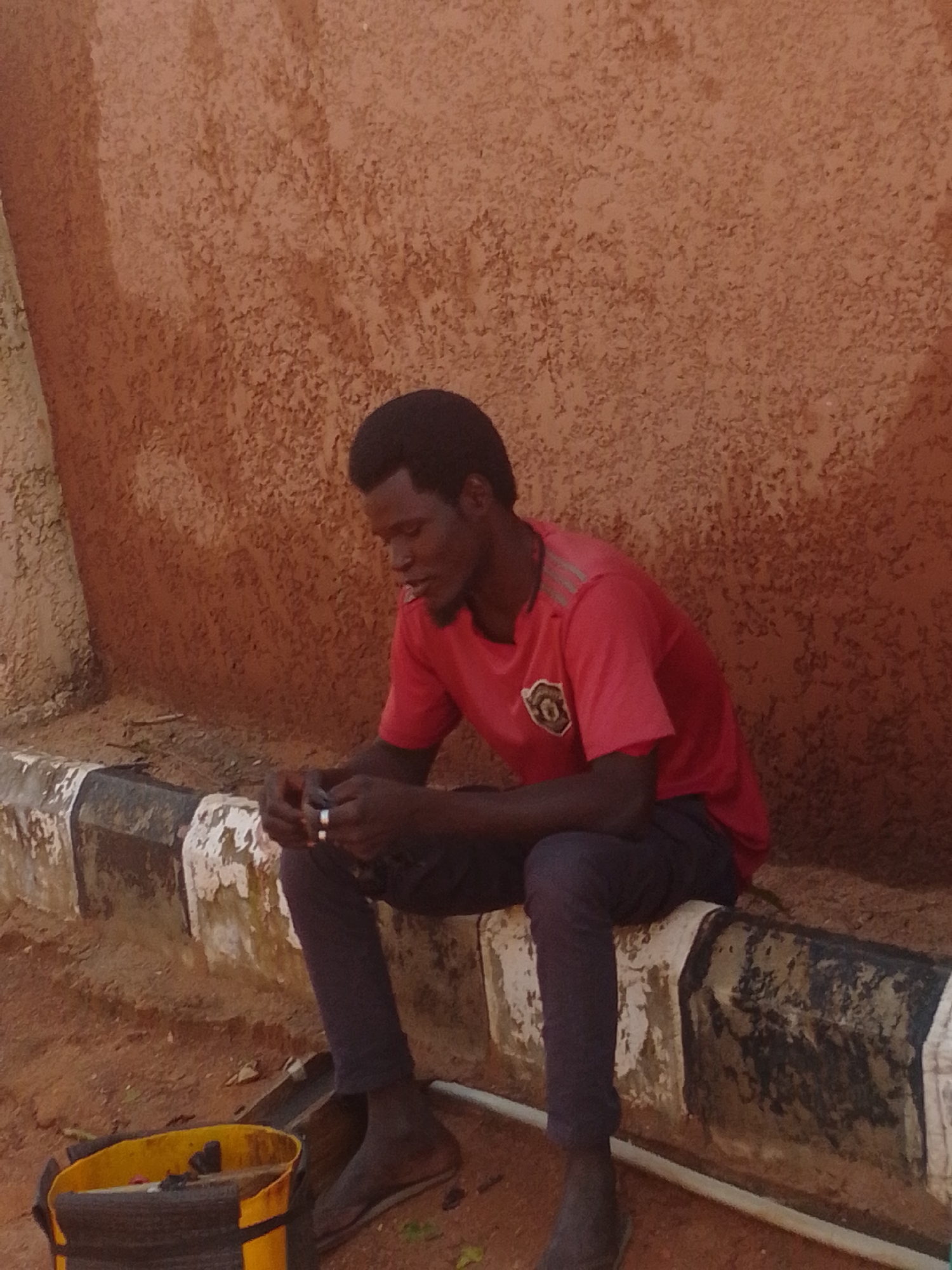 A young Black man with short, dark hair is sitting on a concrete curb in Tempure, Nigeria. He is wearing a red t-shirt with a crest on the left chest and dark gray pants. His hands are clasped in front of him, and he appears to be holding something small, possibly a cigarette, between his fingers. He is looking down and to his right. The curb is painted with alternating black and white stripes. Behind the curb, the ground is sandy dirt. A rough-textured, reddish-brown wall forms the background. To the left of the man, a yellow bucket with black trim sits on the ground. The lighting suggests it is daytime.