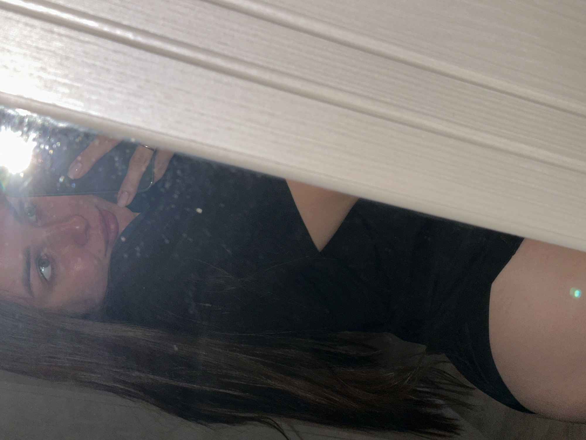 A young woman with fair skin and long, dark hair lies in what appears to be a reflective surface with white slats visible at the top. She is wearing a black top and black bottoms. Her face is turned toward the viewer, and she is holding a cell phone, as if taking a selfie. The location is not discernible from this image.