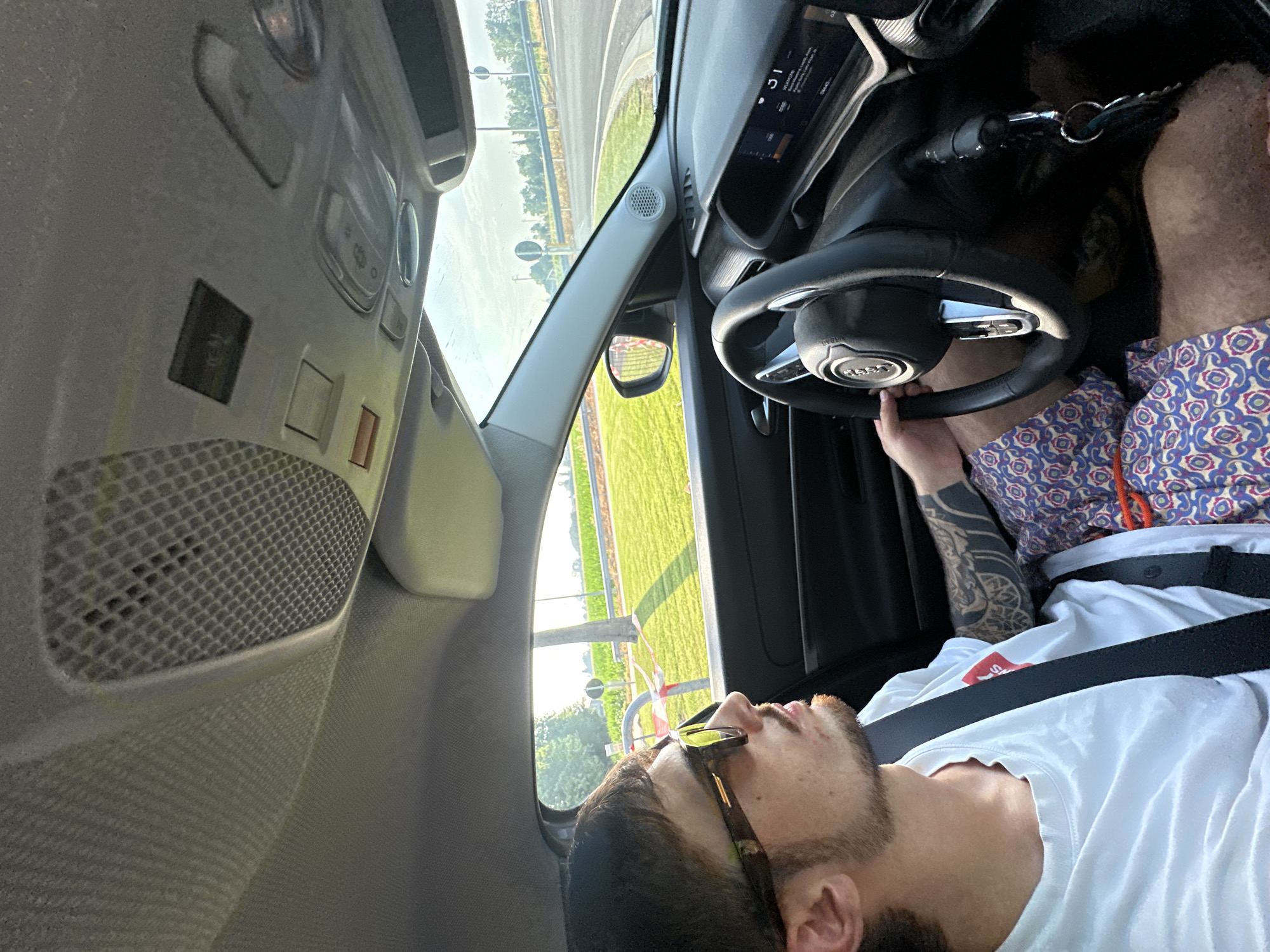 An individual is seated in the driver's seat of a car, looking out the window. The car's interior is visible, including the dashboard, steering wheel, and ceiling console with speakers and lights. The individual is wearing sunglasses, a white t-shirt, and patterned shorts, and has tattoos on their arm. Outside the car, a sunny rural landscape with green fields and trees can be seen. The road appears to be a gentle curve. There is a traffic sign visible in the distance. The location is the city of Chiari, Italy.