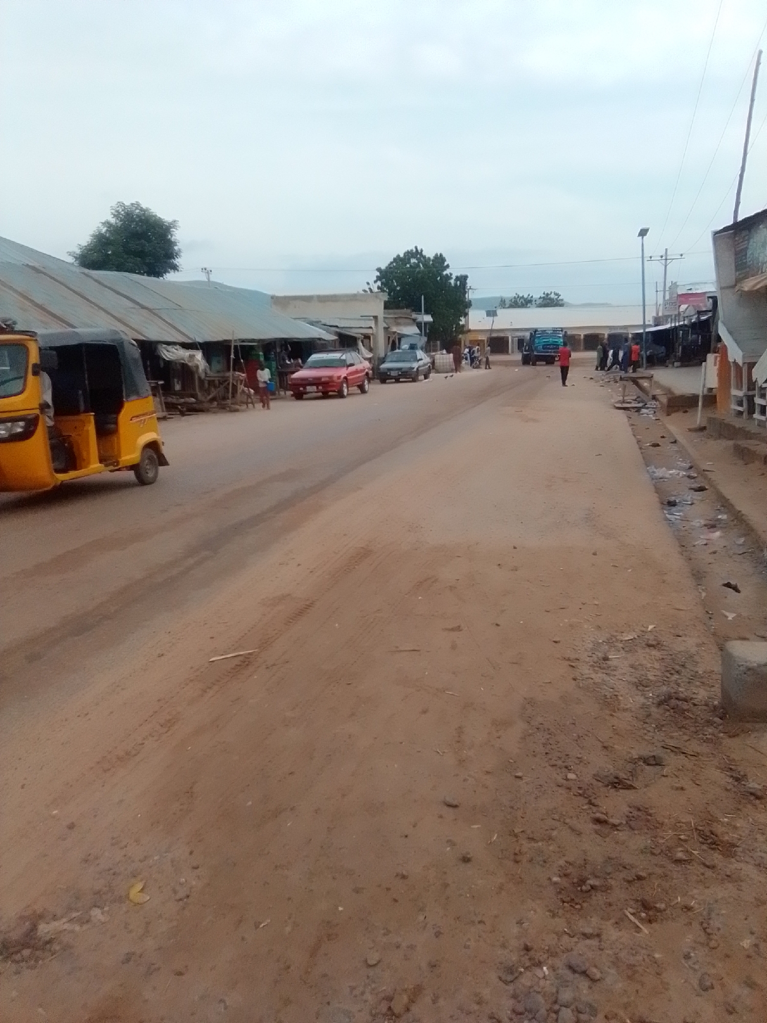 A dirt road in Galadima, Nigeria, is lined with various structures, including corrugated metal-roofed buildings that appear to be shops or stalls. A yellow auto-rickshaw is parked on the left side of the road. Several cars are visible, including a red sedan and a silver sedan further down the road. A blue truck is also present. A few individuals are walking on the road and sidewalks. The environment appears dusty and somewhat unkempt, with debris scattered along the roadside. The sky is overcast.