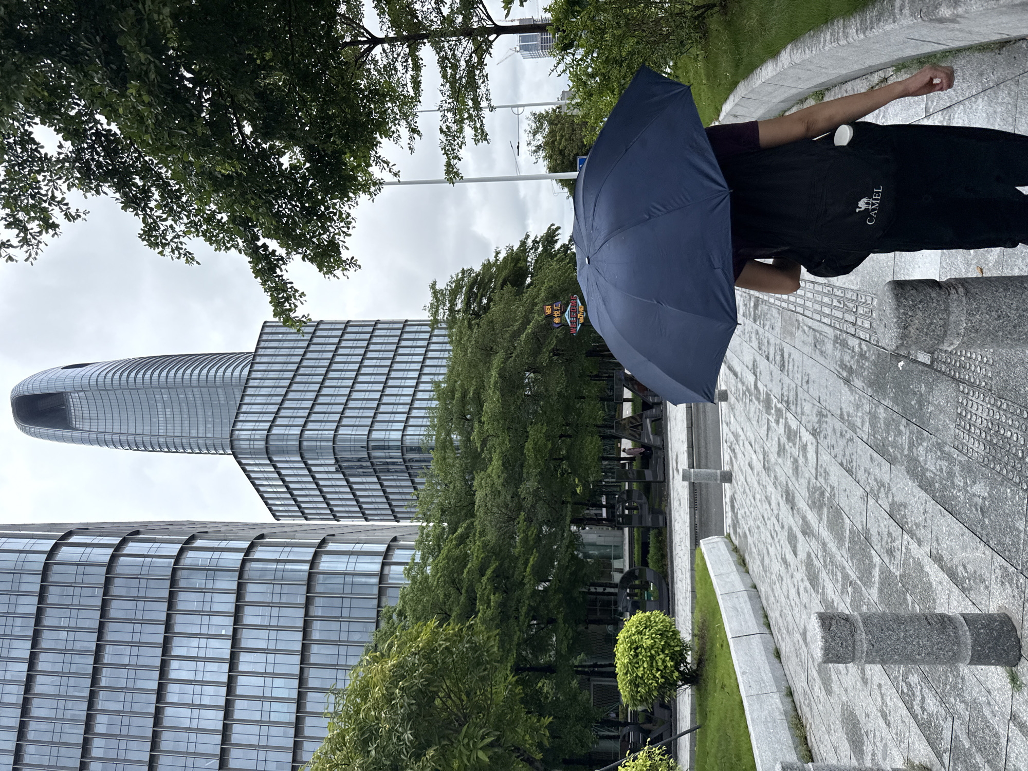 A person walks with a dark blue umbrella open on a wet, paved walkway lined with granite bollards and a low stone wall. The person, seen from behind, wears a dark shirt, black pants, and a black backpack with a white camel logo. The background features modern glass-facade buildings, including a tall, curved skyscraper, and lush green trees. The sky is overcast. The scene appears to be in a city park or plaza area within Haizhu District, China.