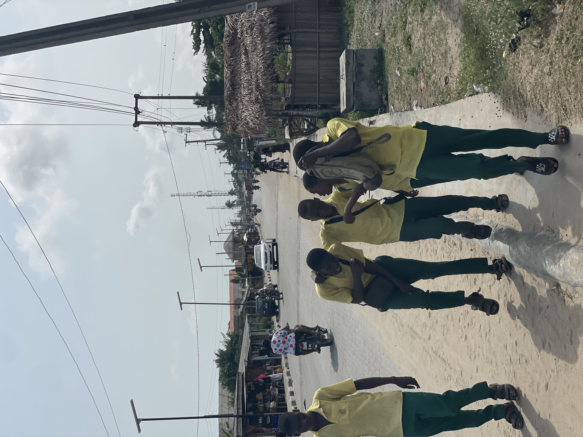 The image shows a street scene in Badagry, Nigeria. Four male figures, presumably students, are walking along a sidewalk adjacent to a road. They are wearing a uniform of green pants and light yellow shirts. Each of the boys is carrying a bag. The road has light-colored asphalt and shows vehicular traffic, including cars and motorcycles, moving in both directions. Buildings, some with thatched roofs, border the road. Power lines and utility poles are visible above the street. The weather appears sunny and the sky is partly cloudy.