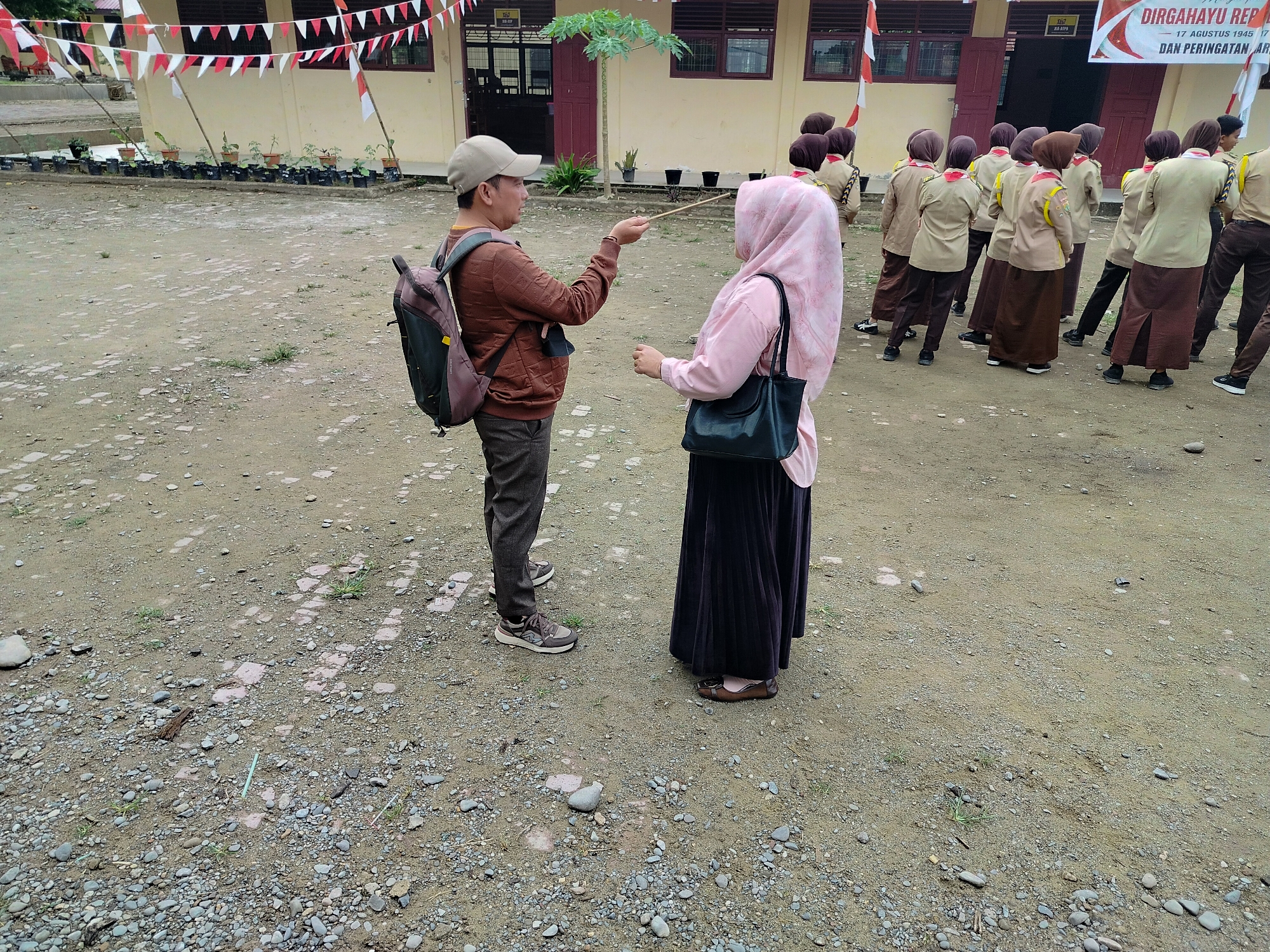 In Lawebekung, Indonesia, a male figure wearing a brown sweatshirt, grey pants, a beige cap, and a backpack stands facing a female figure wearing a pink hijab, a pink long-sleeved shirt, and a dark skirt. The man holds a thin stick in his right hand, pointing it towards the woman. The woman has her left hand extended towards the stick, and she carries a dark handbag. Behind them, a group of individuals, likely students, are dressed in matching tan uniforms with brown skirts and headscarves, forming a line and facing away from the camera. They are positioned in front of a light-colored building adorned with red and white triangular flags. The ground is a mixture of dirt and small stones.