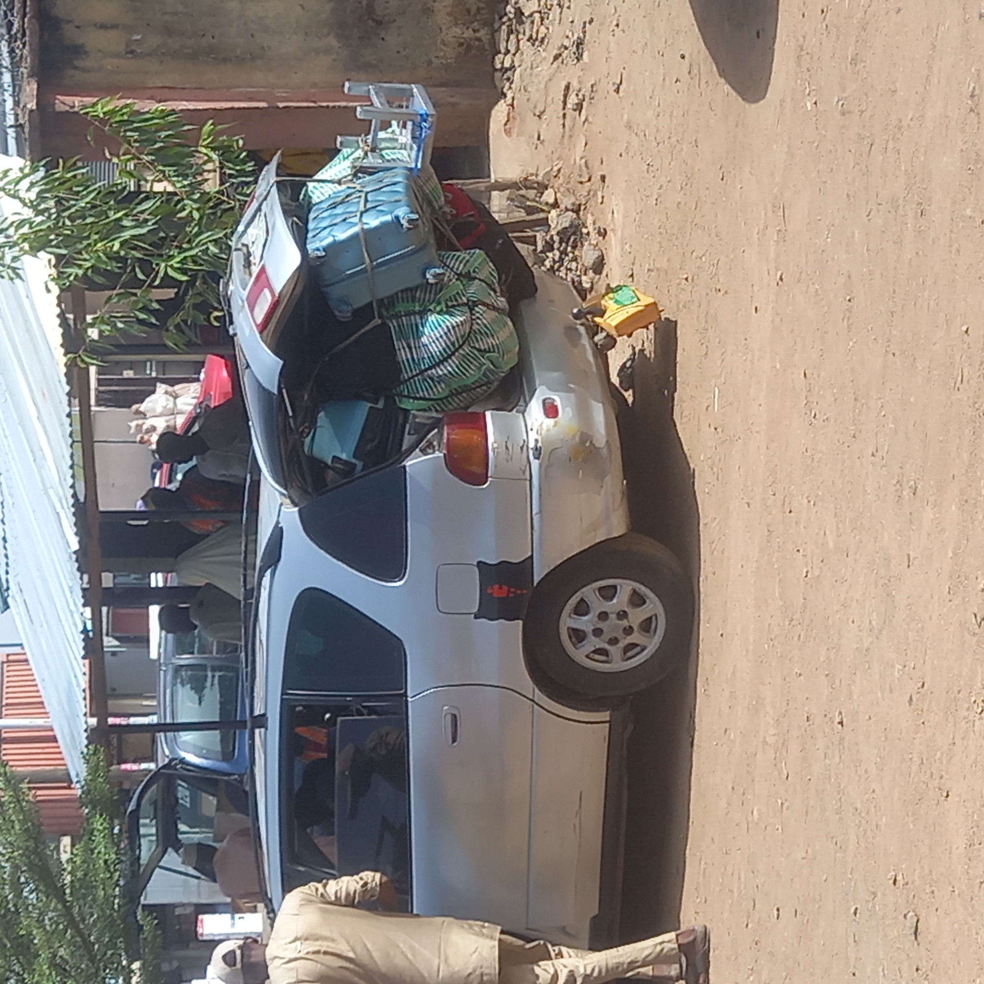 The image, rotated counter-clockwise, captures a silver station wagon or SUV parked on an unpaved, dusty ground. The vehicle's trunk is open and heavily laden with luggage, including at least two blue suitcases and multiple striped cloth bags, all appearing to be tied down. A ladder or roof rack is secured on top of the stacked items. The car shows visible signs of wear, with scratches and dents on its body.
Several people are present in the scene. One individual, dressed in a light brown or tan long-sleeved outfit, stands near the front driver's side of the car, facing away from the camera. Other figures are partially visible inside and around the vehicle, their features indistinct. To the left of the car, a building with a corrugated metal roof is partially seen, alongside a tree or large bush with green foliage. The ground is light brown and appears to be dirt or sand. A small, yellow object, possibly a toy truck or container with a green top, rests on the ground near the car's rear wheel, displaying a faint "C" mark. The scene is brightly lit by daylight, suggesting a sunny day. The overloaded car points to an event of travel or transport of goods, possibly in Sardauna, Nigeria.