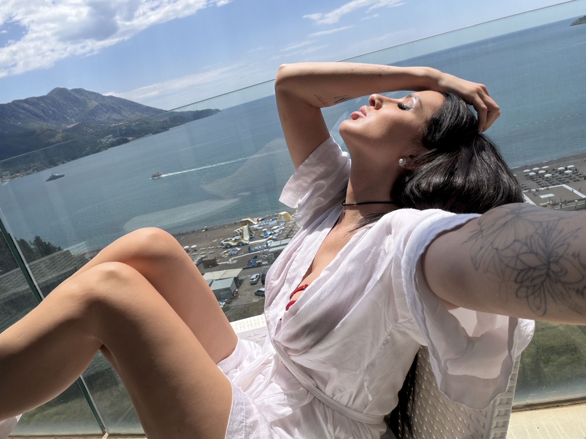 A woman wearing a white dress and visible red swimwear underneath sits on a balcony with a clear glass railing. She has dark hair, fair skin, and tattoos on her arms. Her head is tilted back and her hand is in her hair, suggesting she is basking in the sun. Behind her is a view of the city of Boreti, Montenegro. The background includes a mountain, a body of water with boats, a beach with umbrellas and beachgoers, and various buildings. The sky is partially cloudy.