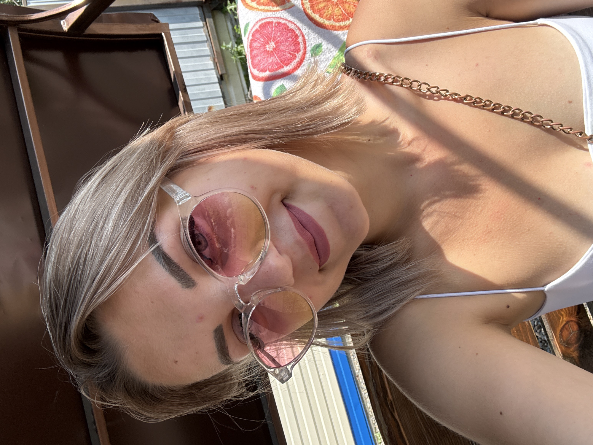The image is a close-up selfie of a young woman with blonde hair and sunglasses, outdoors in Krasnogorskoye, Russia. She is wearing a white tank top with a gold chain strap. The background shows a sunny day with a fence and some greenery. The overall scene suggests a casual, outdoor setting.