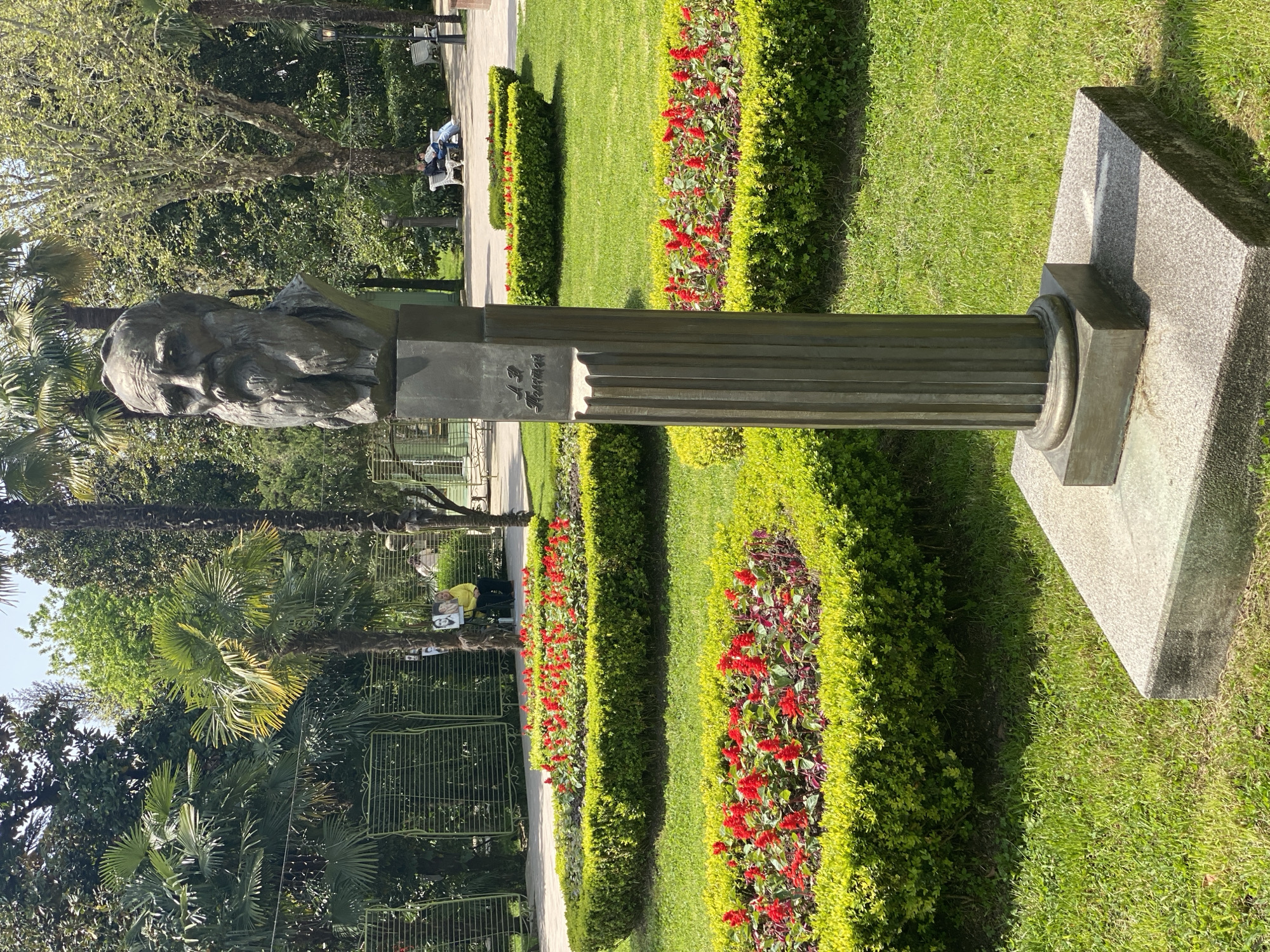 The image features a bronze bust of an older man with a distinguished beard and a serious expression, positioned prominently in a sunny park. The bust is supported by a fluted, dark metallic column, which rests on a rectangular, two-tiered granite pedestal. On the column, below the bust, the Cyrillic inscription "Л. Н. Толстой" (L. N. Tolstoy) is clearly visible.
The monument is set amidst a manicured garden landscape. Two parallel rows of neatly trimmed green hedges run across the midground, with a vibrant display of red flowers planted between them. Beyond these hedges, there are lush green lawns and paved pathways. In the background, various trees, including tall palm trees and deciduous trees with abundant green foliage, create a natural backdrop.
The scene is brightly lit by the sun, casting distinct shadows and suggesting a clear, sunny day, likely in the late morning or early afternoon. In the upper right background, two people are visible sitting on a park bench, seemingly relaxing or enjoying the park environment. A green metal fence is also present in the background. The setting is a well-maintained public park in Sochi, Russia, offering a peaceful atmosphere.