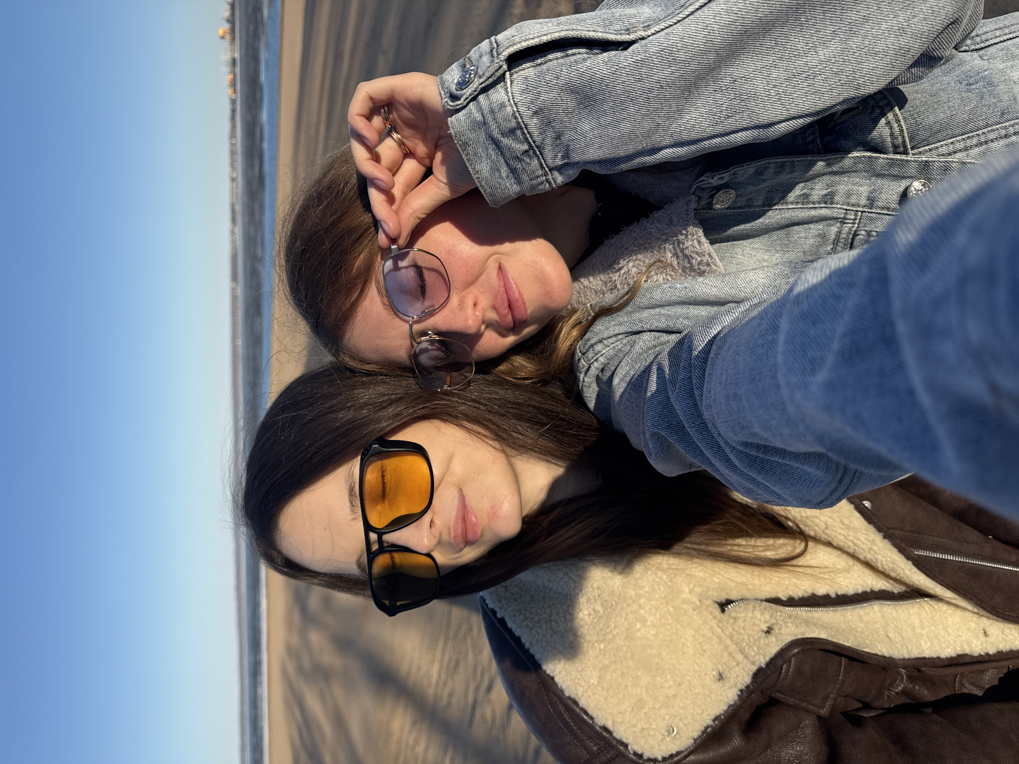 Two young women lie on a sandy beach. One woman is wearing a denim jacket, and the other is wearing a brown leather jacket with a shearling collar. Both women are wearing sunglasses. The ocean and sky are visible in the background. The location is identified as a beach in the city of Noviy, Russia.