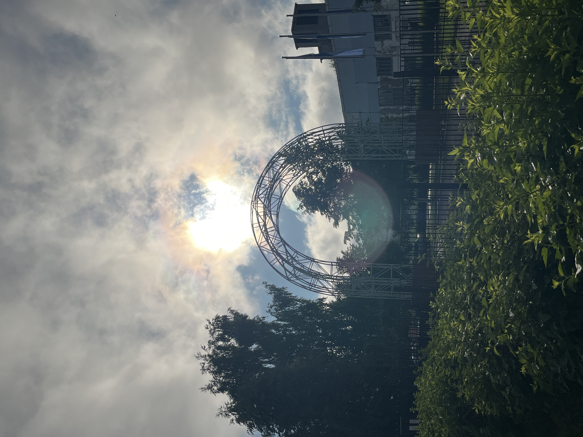 The image depicts an outdoor scene on a cloudy day, with the sun partially visible and creating a halo effect. A large, metallic arched structure, possibly a stage prop or an architectural element, dominates the center of the frame. It is supported by a scaffolding-like base. To the left of the arch, a tall, leafy tree stands prominently. In the background, a building with a light-colored facade and multiple windows is visible, along with three flags on poles. In front of the arch and building, there is a black metal fence. A dense line of green bushes runs along the bottom of the image, in front of the fence. The overall impression is that of an outdoor public space or park area. The context provided indicates the location is in the city of Chaikovsky, Russia.