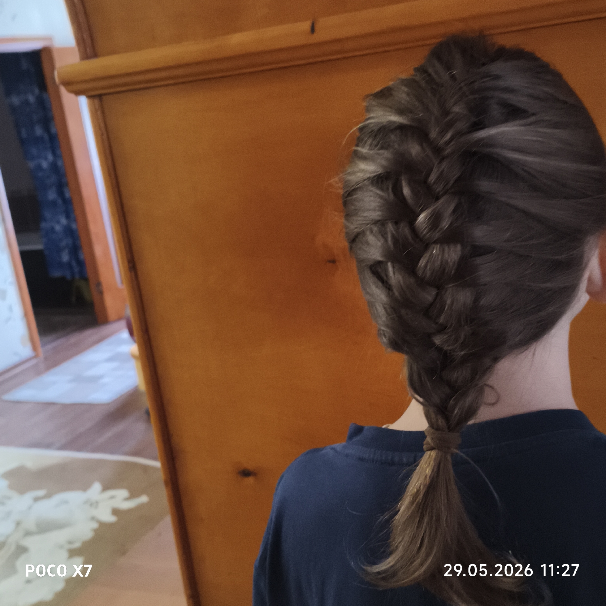 A view of a person's back with their head and shoulders. The person has dark brown hair braided in a French braid style, ending in a ponytail. The person is wearing a navy blue shirt. Behind the person is a large wooden cabinet or wardrobe. The background shows a blurred view of a room with a doorway and a patterned rug on the floor. The photo was taken with a POCO X7 camera. The location is likely an interior space, potentially a home. There is no information in the image to indicate that it was taken in the city of Chaikovsky, Russia.