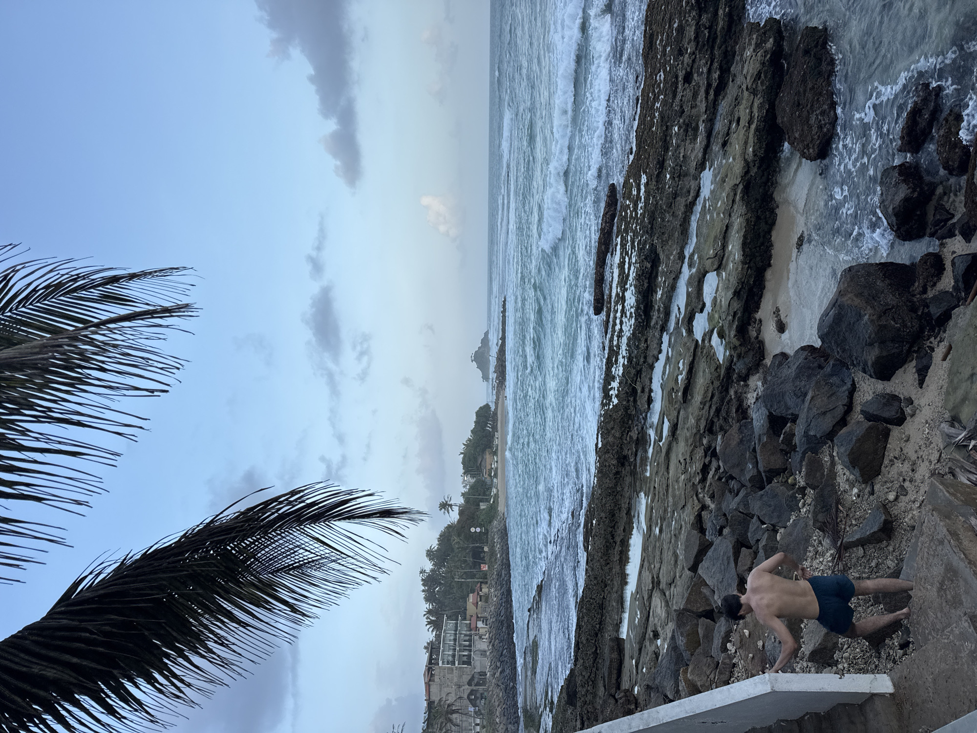 This is a landscape photograph taken from a rocky shoreline looking out at the ocean under a cloudy sky. The foreground features a rough, rocky beach with some tide pools and a concrete staircase with a white railing on the left. A young man with dark hair, wearing dark blue swim trunks, is ascending the staircase from the beach. Behind him, the rocky shore transitions to a sandy beach with some vegetation and buildings, including a multi-story structure with balconies, and palm trees lining the coast. The ocean is a vibrant blue with moderate waves breaking on the shore. In the distance, a small island with vegetation and a structure on top is visible. A frond of a palm tree hangs down from the top left corner of the frame, partially obscuring the sky. The lighting suggests it is either early morning or late evening.