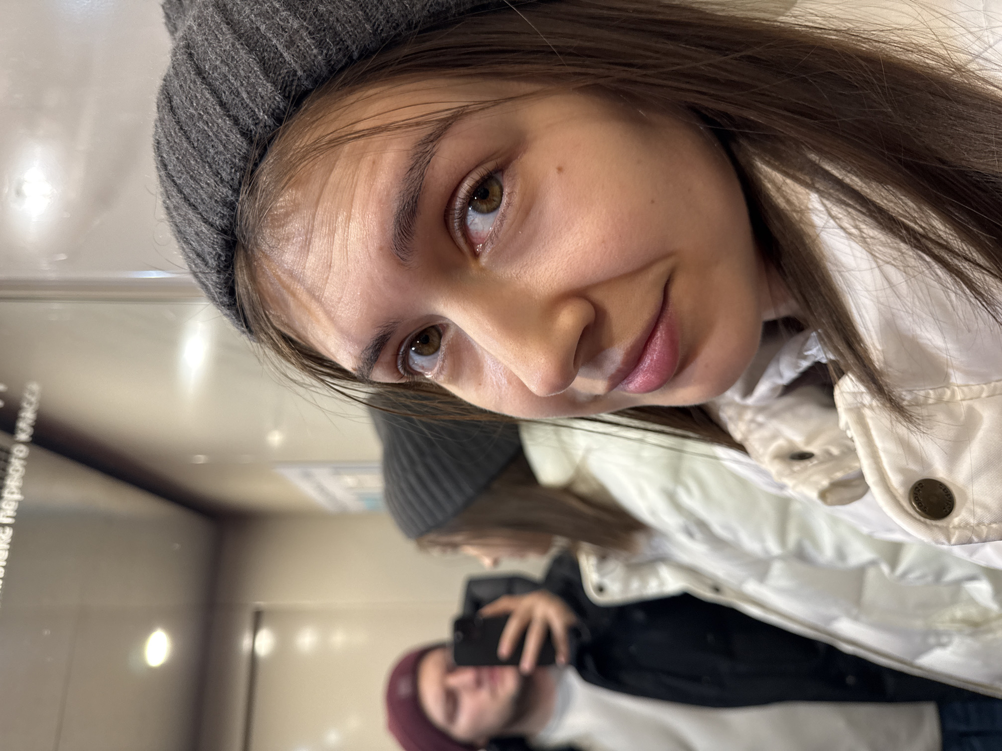 A young woman with light brown hair and brown eyes wearing a gray beanie and a white puffy jacket is taking a selfie in an elevator. The elevator's mirrored wall reflects the woman and a blurry image of a man in a maroon beanie holding a smartphone. A sign with Cyrillic text "в потоке первого класса" is visible on the left side of the elevator. The location is likely Krasnoyarsk, Russia, based on the Cyrillic text on the sign.